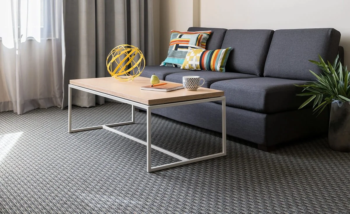 Living room with a grey sofa, patterned cushions, a wooden coffee table with a decorative yellow sphere, a book, a notebook, a pear, a mug, a small potted plant, and curtains by a window.