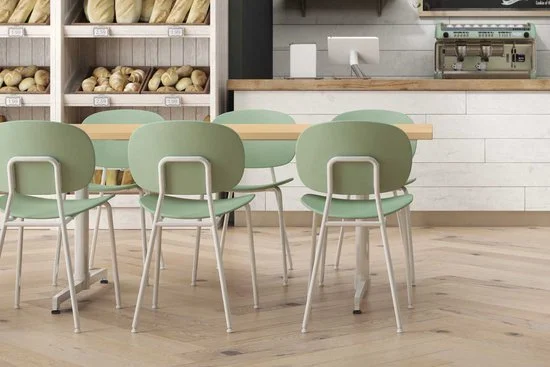 Empty green chairs around a wooden table in a bakery or cafe with shelves of bread and a coffee counter in the background.