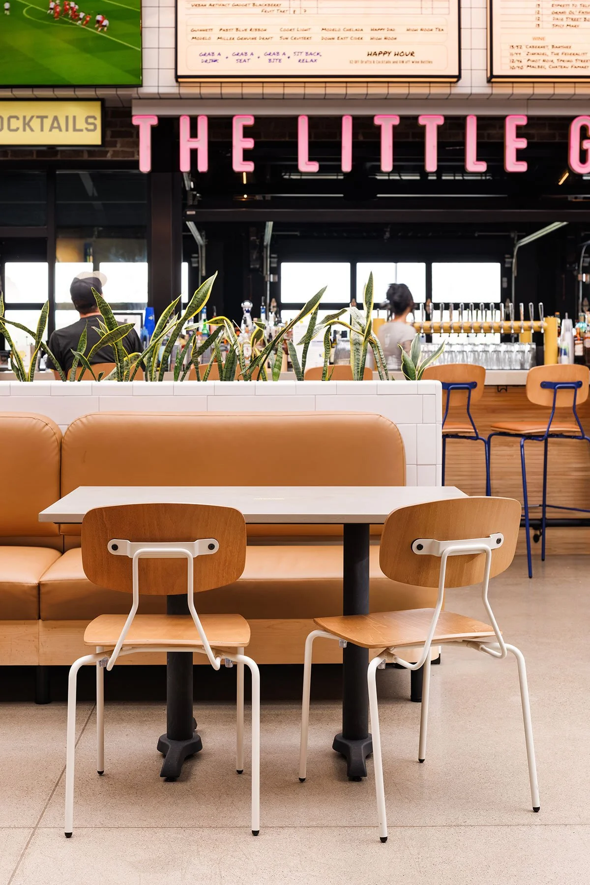 Interior of a restaurant or bar with a table and two chairs in the foreground, a booth with plants behind it, and a bar with stools and bottles in the background. The sign 'THE LITTLE G' is partially visible, and a menu or screen is displayed above.