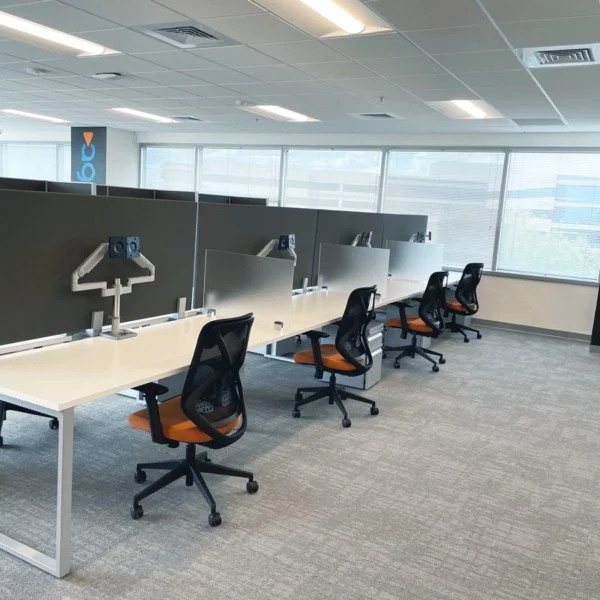Four workstations with black office chairs and dividing cubicle panels in a modern office space with large windows.