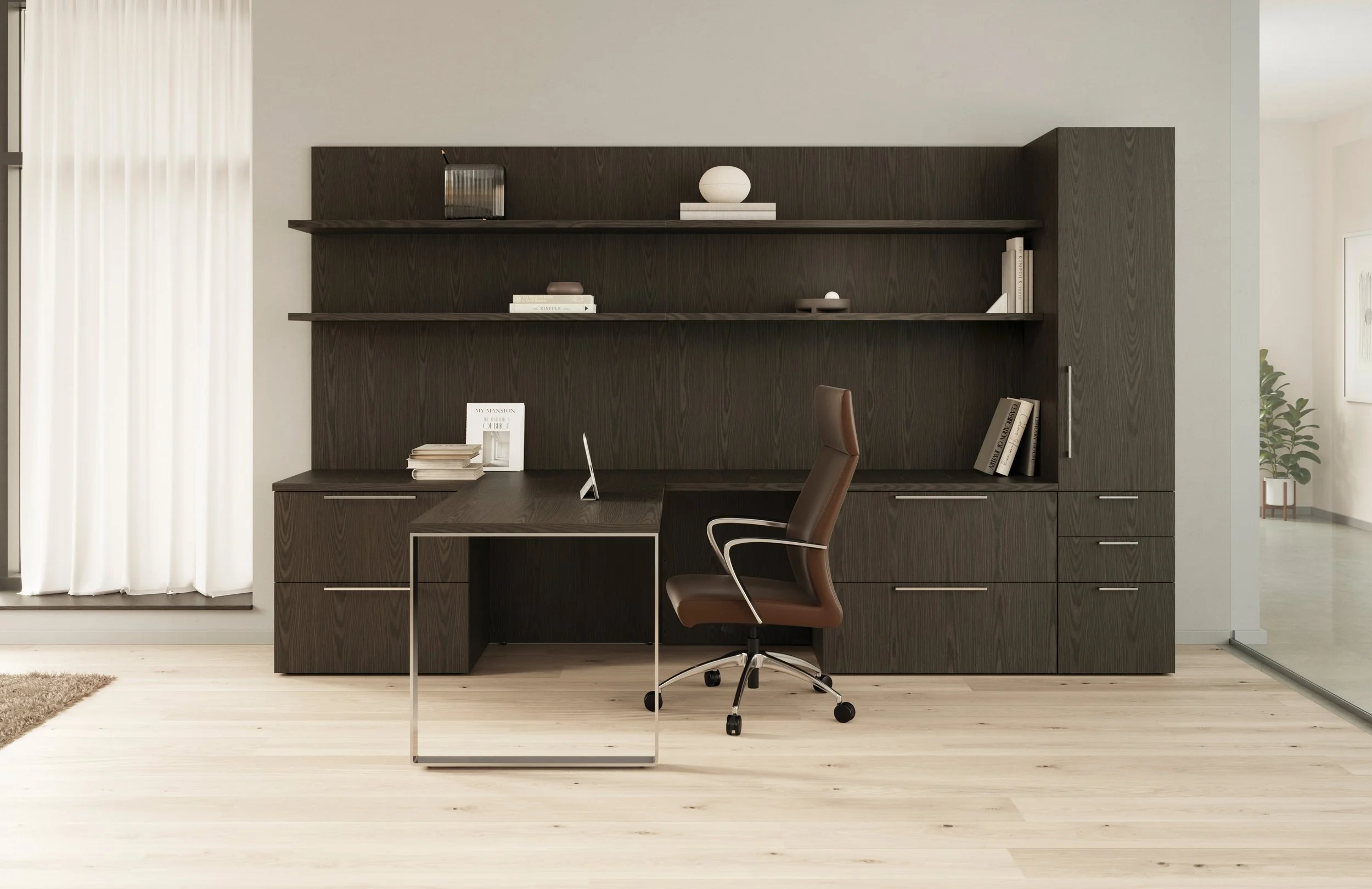 Home office with dark wood desk, brown office chair, and built-in dark wood shelving unit with books and decor, near window with white curtains on light wood flooring.