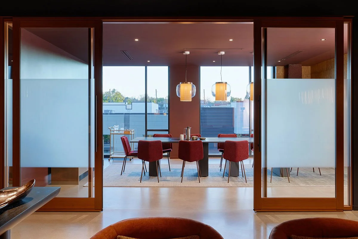 Modern dining room seen through glass sliding doors, featuring a round table with red chairs, pendant lights, large windows with a city view, with a beige rug and minimal decor.