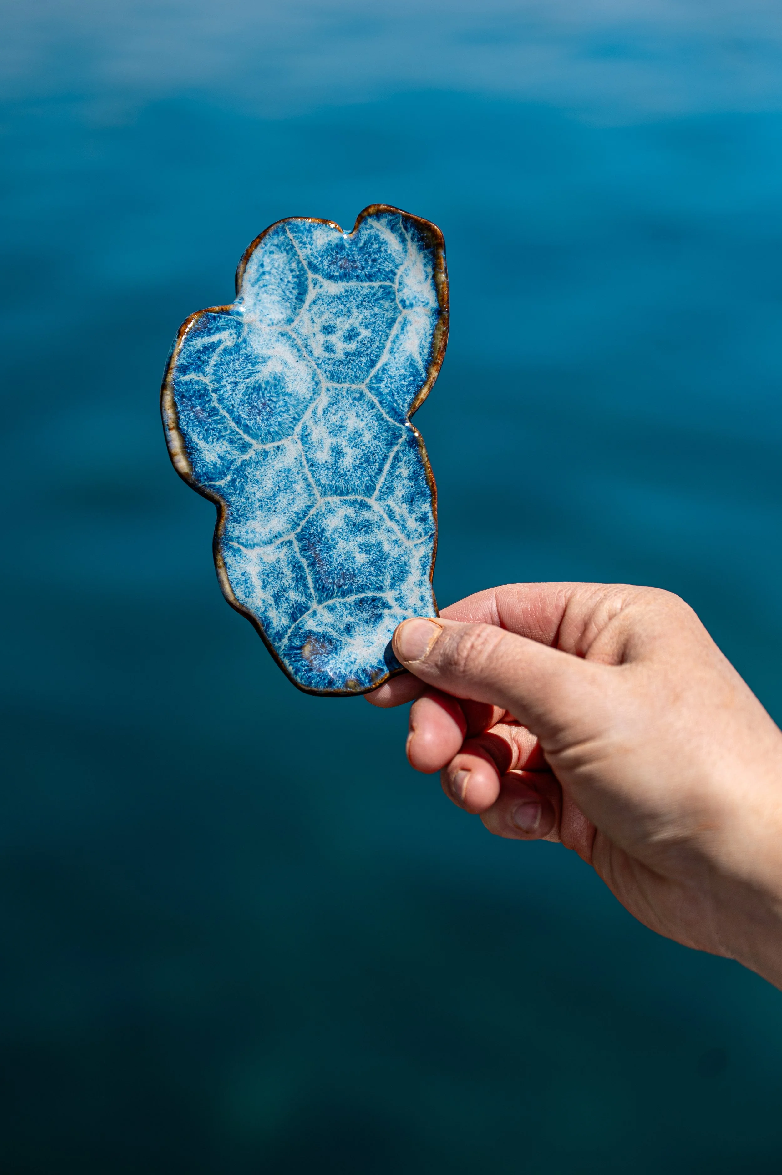 A hand holding a uniquely shaped piece of blue sea glass against a blue background.