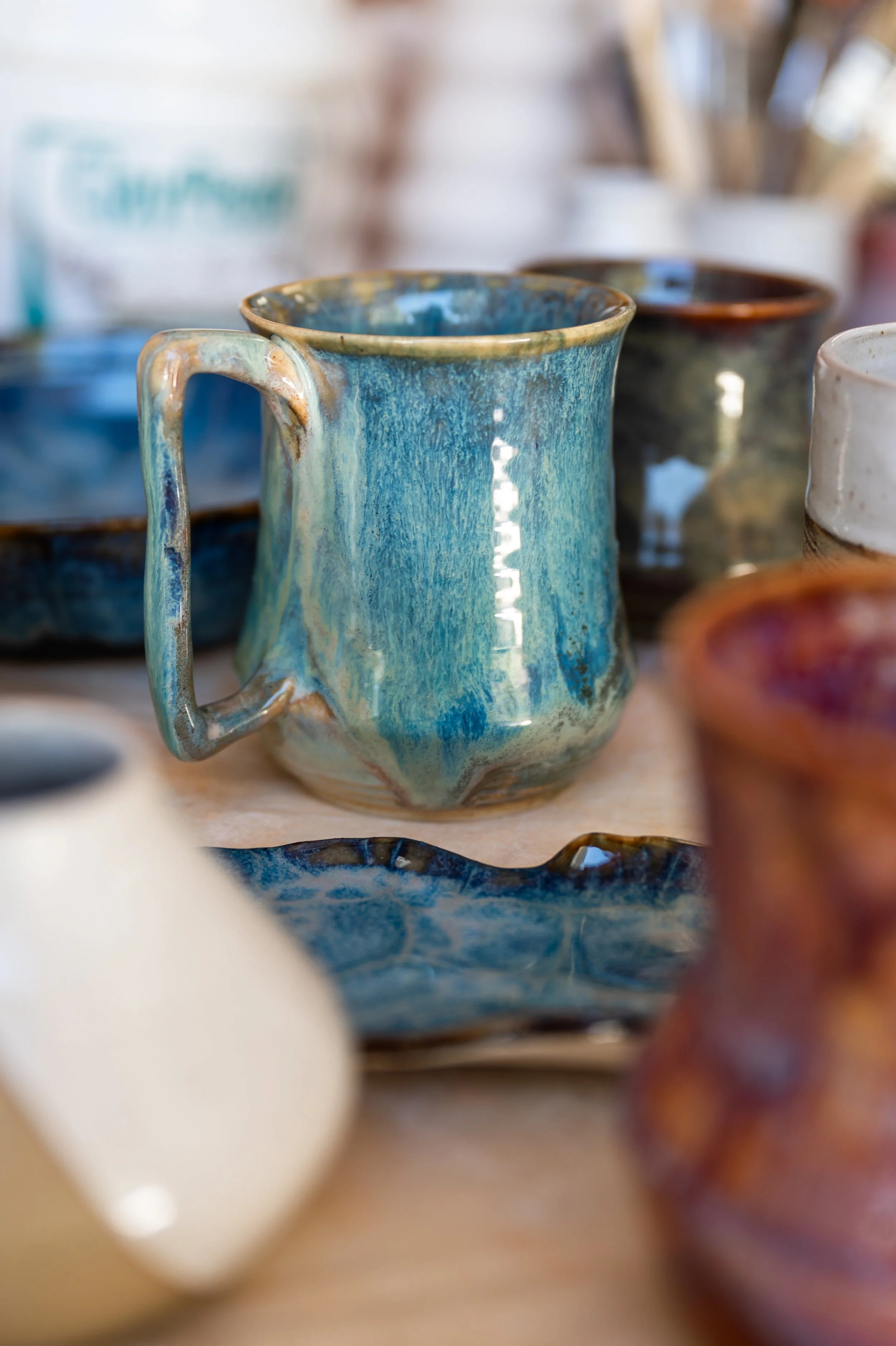 Various handmade ceramic mugs displayed on a table, with vibrant blue, white, and earth-toned glazes, showcasing different shapes and glaze effects.