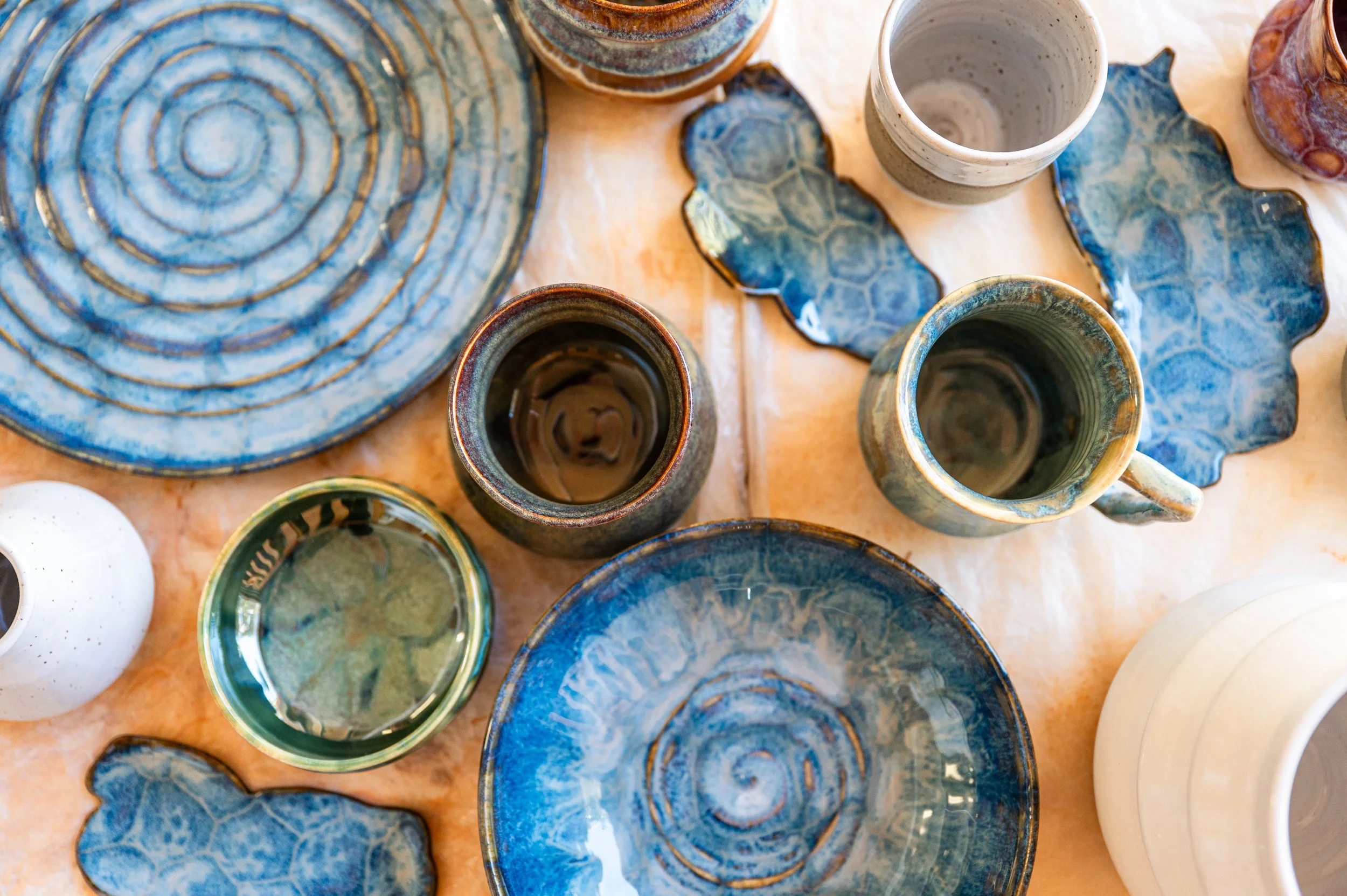 An assortment of handmade pottery cups, plates, and bowls in various earth-tone colors and textures displayed on a wooden surface.