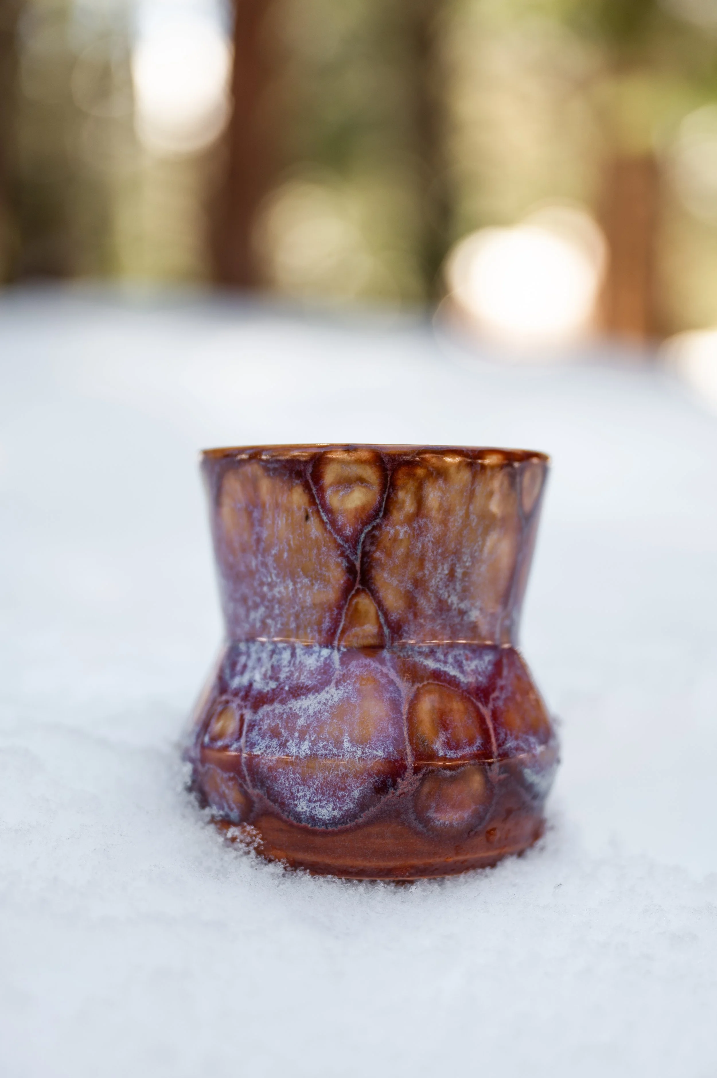 Close-up of a small ceramic or clay cup with a textured, glossy brown and orange glaze, set on snow with a blurred background of trees and sunlight.