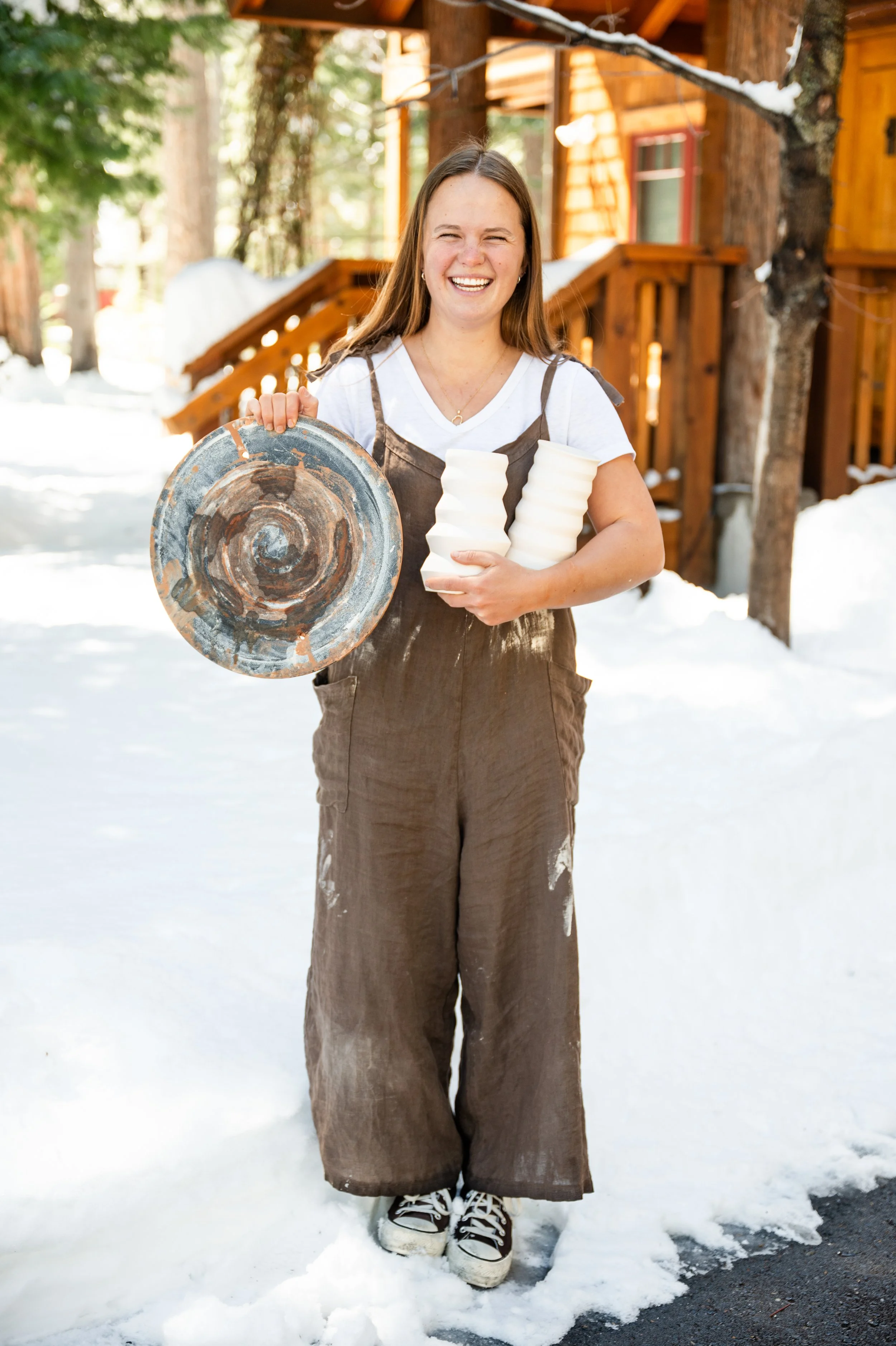 A young woman with long brown hair smiling in outdoor snowy setting, standing in front of a wooden cabin, wearing a white T-shirt, brown overalls, and black sneakers, holding a metal pan and foam cups.