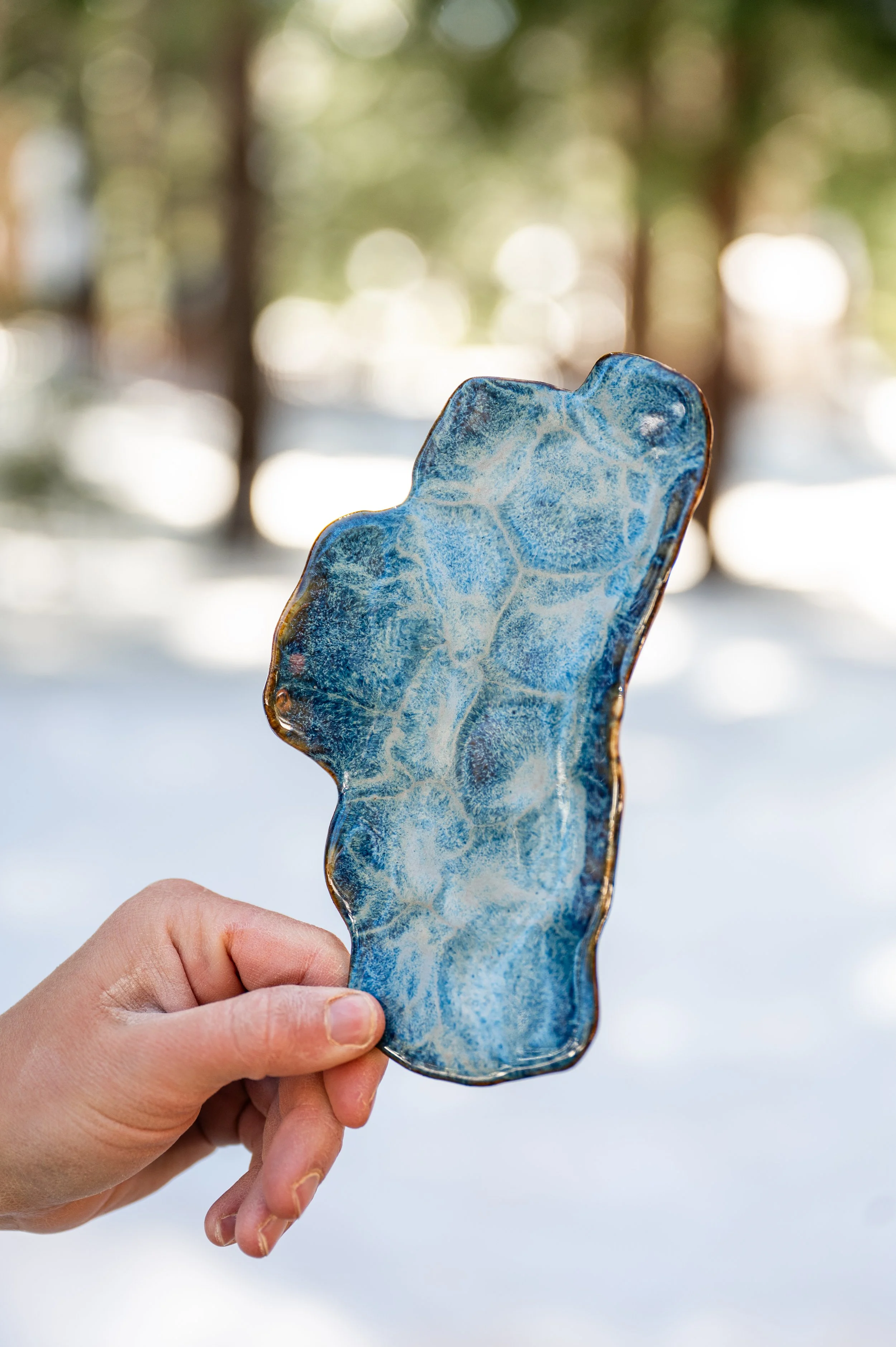 A close-up of a person's hand holding a piece of blue and clear textured glass with a wavy shape, against a blurred outdoor background with trees and snow.