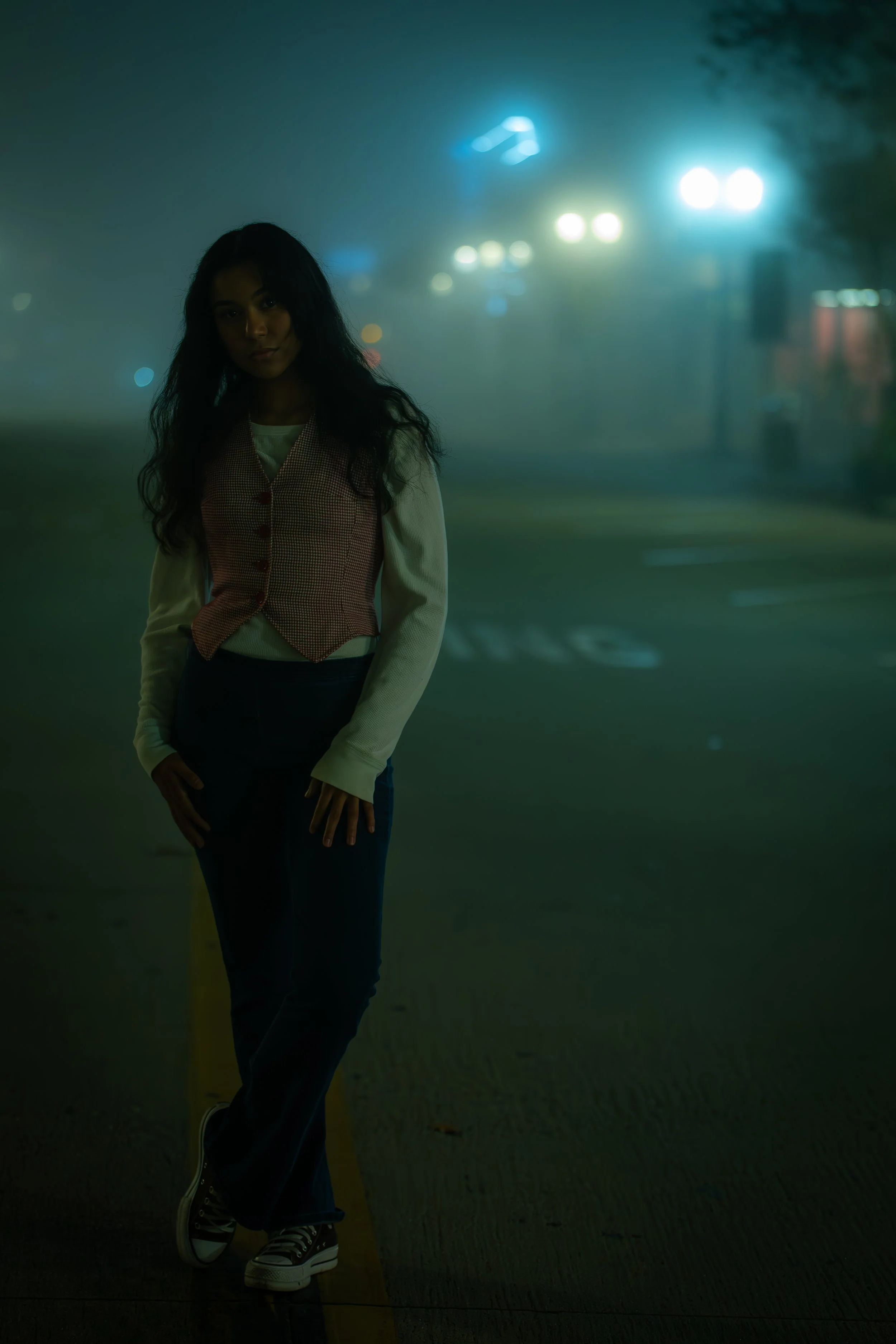 A woman with long dark hair standing on a foggy street at night, illuminated by blue streetlights, wearing a beige sweater, checkered vest, dark pants, and sneakers.