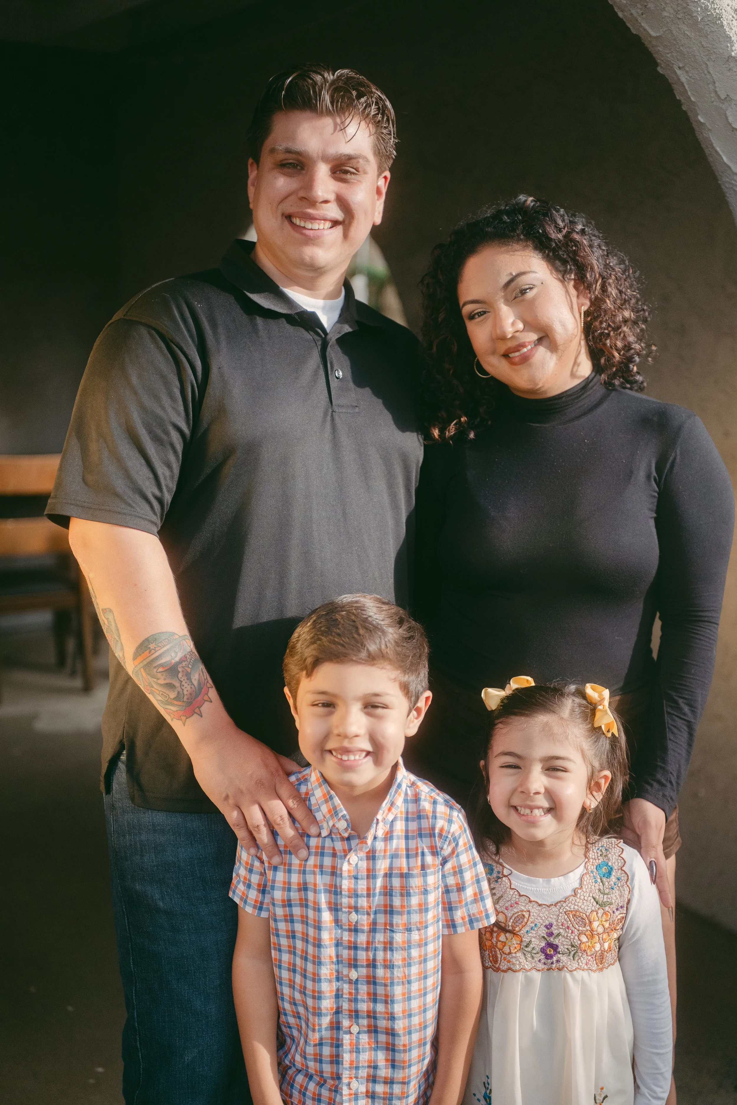 A smiling family of four posing together indoors, including a man, a woman, a young boy, and a young girl.