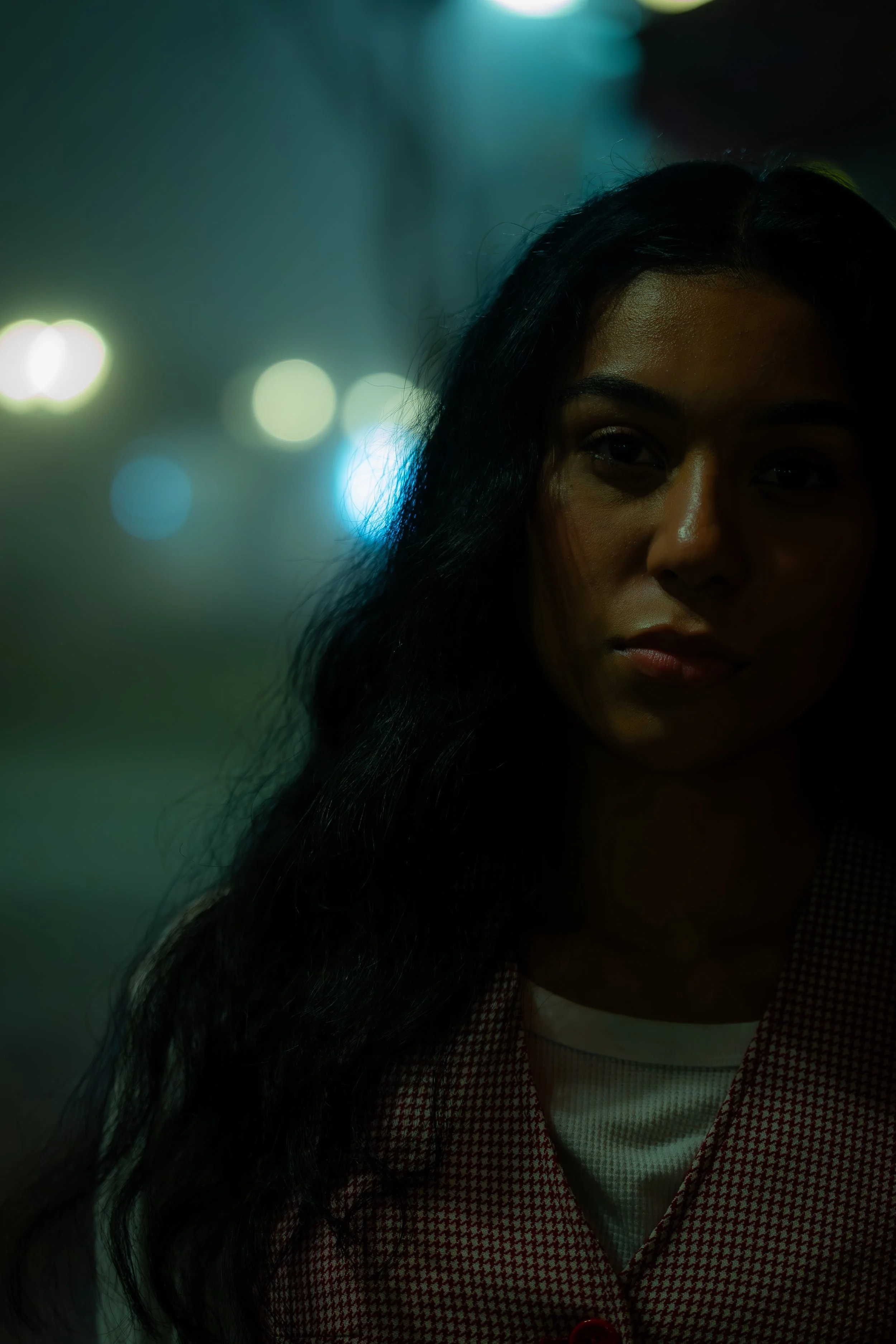 A young woman with long dark hair standing in a dimly lit urban setting at night, with blurred colorful city lights in the background.