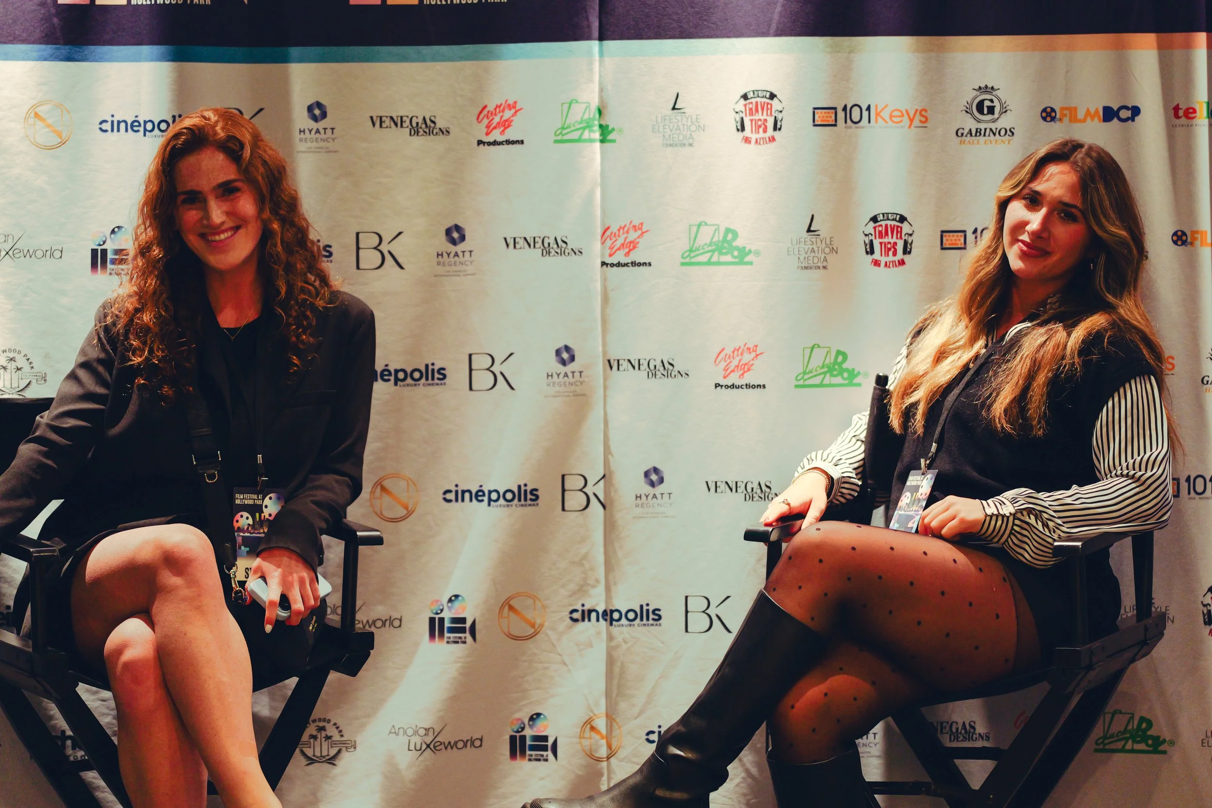 Two women sitting on director's chairs in front of a backdrop with logos at an event. The woman on the left has curly red hair, wearing a black blazer and skirt. The woman on the right has long blonde hair, wearing a black vest over a striped shirt w