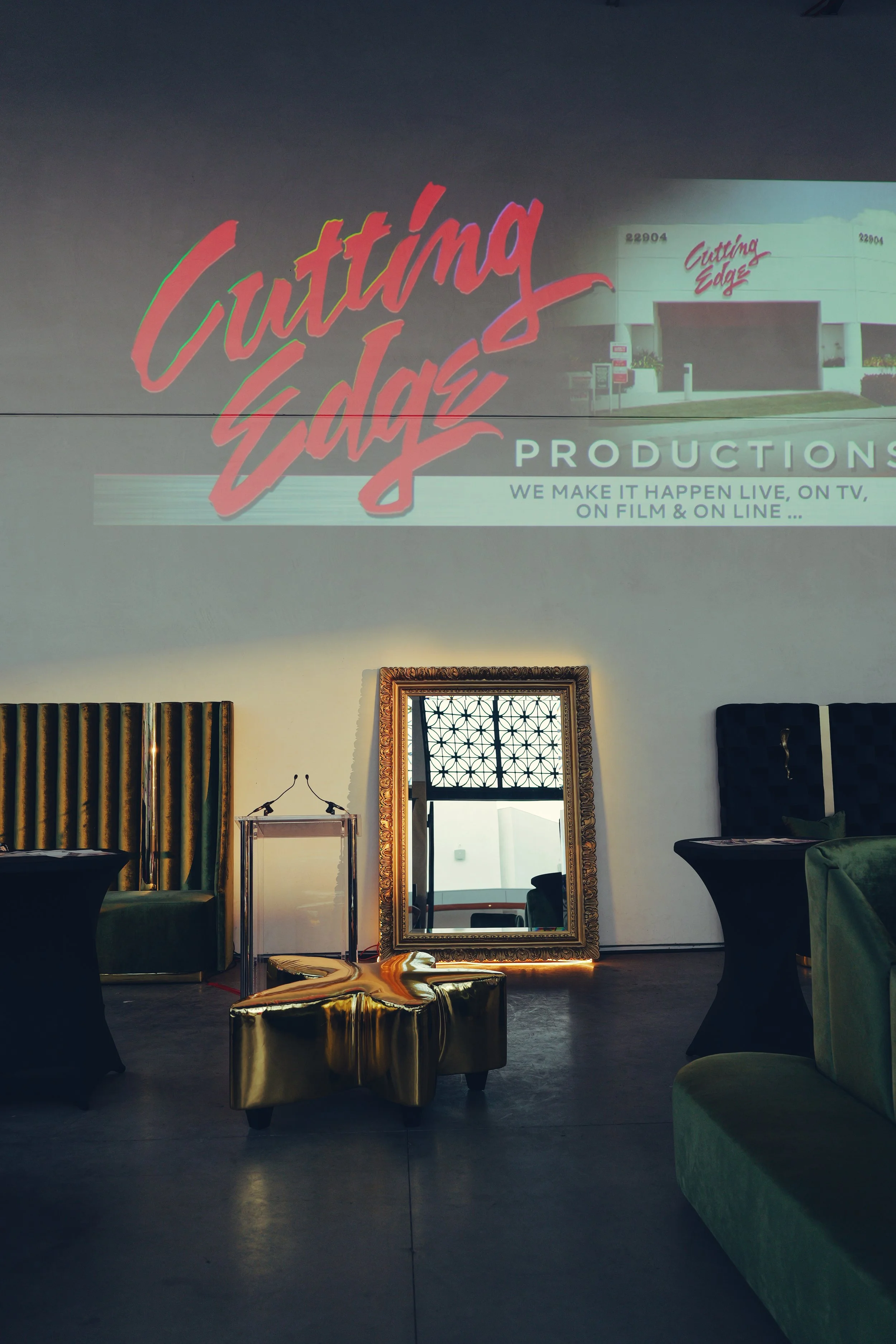 Interior of a room with a large framed mirror, green upholstered chairs, and a gold Starbucks star-shaped table, with a projection on the wall displaying 'Cutting Edge Productions' with an image of a building.