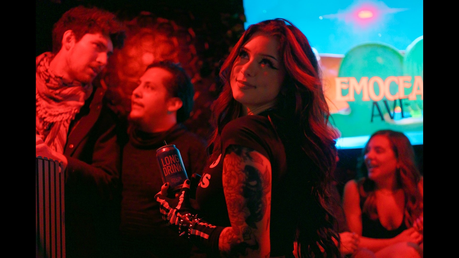 A group of people at a social event, with a woman with red hair and tattoos in the foreground looking to the side, holding a can of long drink, surrounded by others smiling and chatting, under colorful lighting with a sign in the background that read