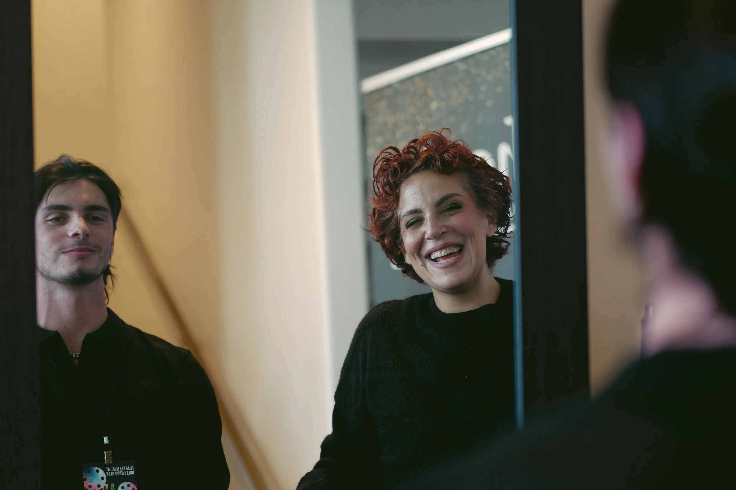 Two people smiling and looking at each other in an elevator mirror, woman with curly short red hair and black sweater, young man with dark hair and black shirt, with an elevator control panel visible.