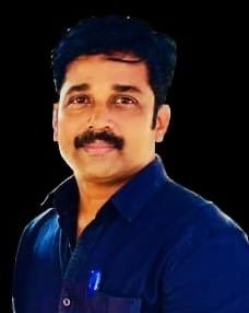 A man with dark, curly hair, a mustache, wearing a dark blue shirt, smiling slightly, against a plain black background.