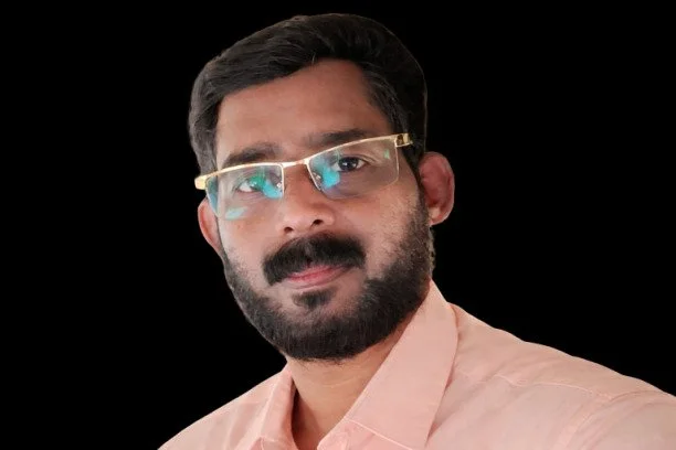 Portrait of a man with glasses and a beard, wearing a light pink shirt, against a black background.