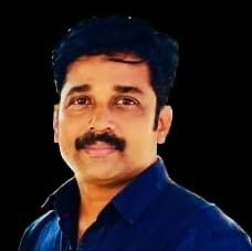 A man with dark hair and a mustache, wearing a dark blue shirt, posing against a dark background.