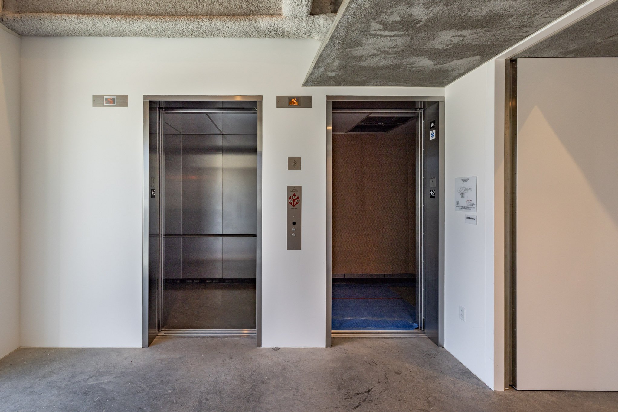 Two elevator doors, one open and one closed, in a modern building. The open elevator shows a brown interior; the closed one has a metallic surface. There are buttons and signage on the white wall between them.