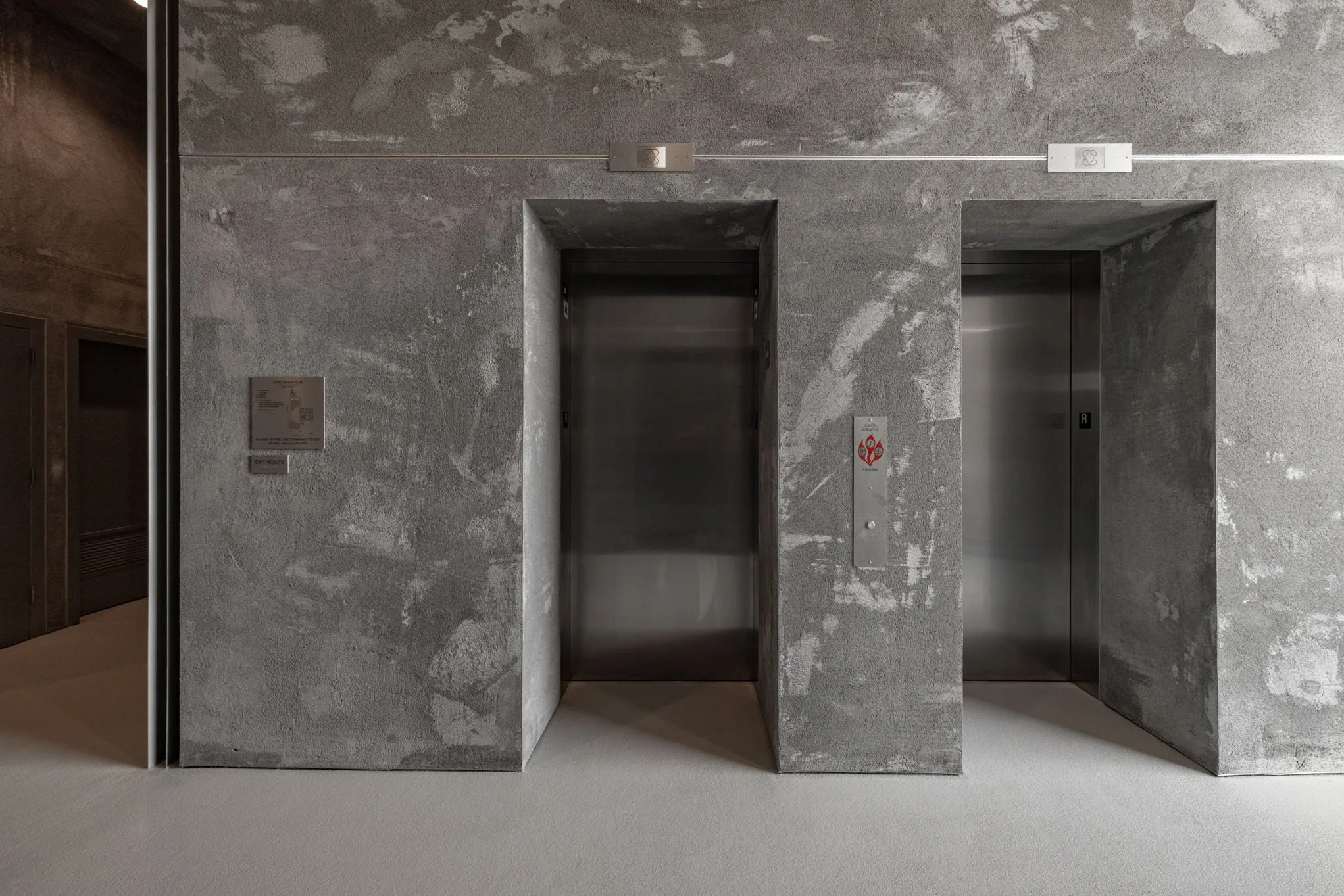 Two elevator doors set in a gray, textured concrete wall with minimal signage.