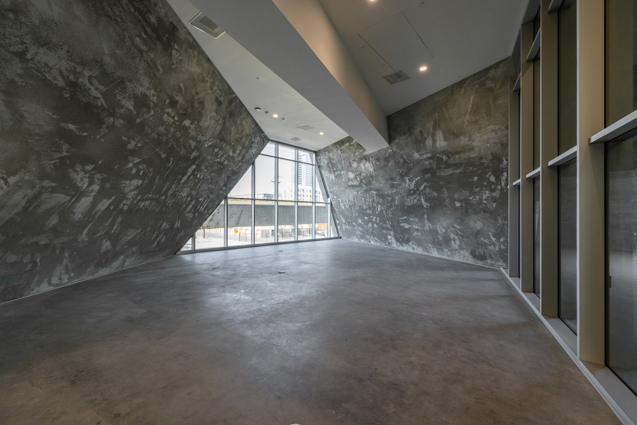 Empty modern interior space with concrete walls, a polished concrete floor, large floor-to-ceiling windows, and ceiling lights.