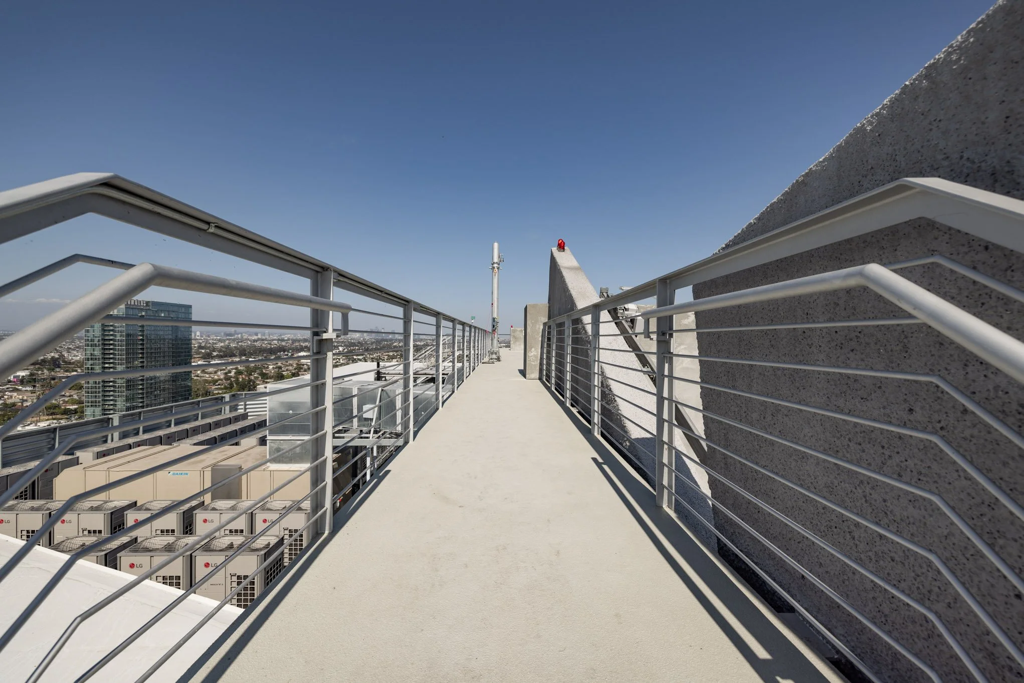 Rooftop walkway with metal railings on a high building with a view of the cityscape, blue sky, and large concrete structures.