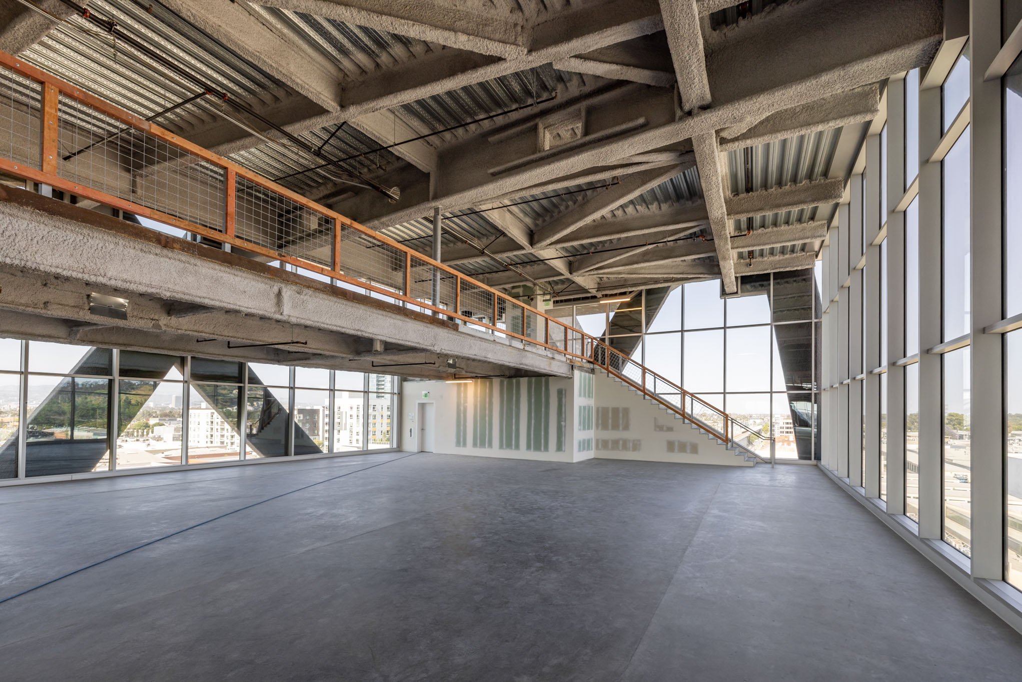 Interior of a modern, unfinished commercial space with large windows, concrete flooring, and an exposed ceiling with beams and insulation. There is a staircase leading to an upper level with a safety railing.