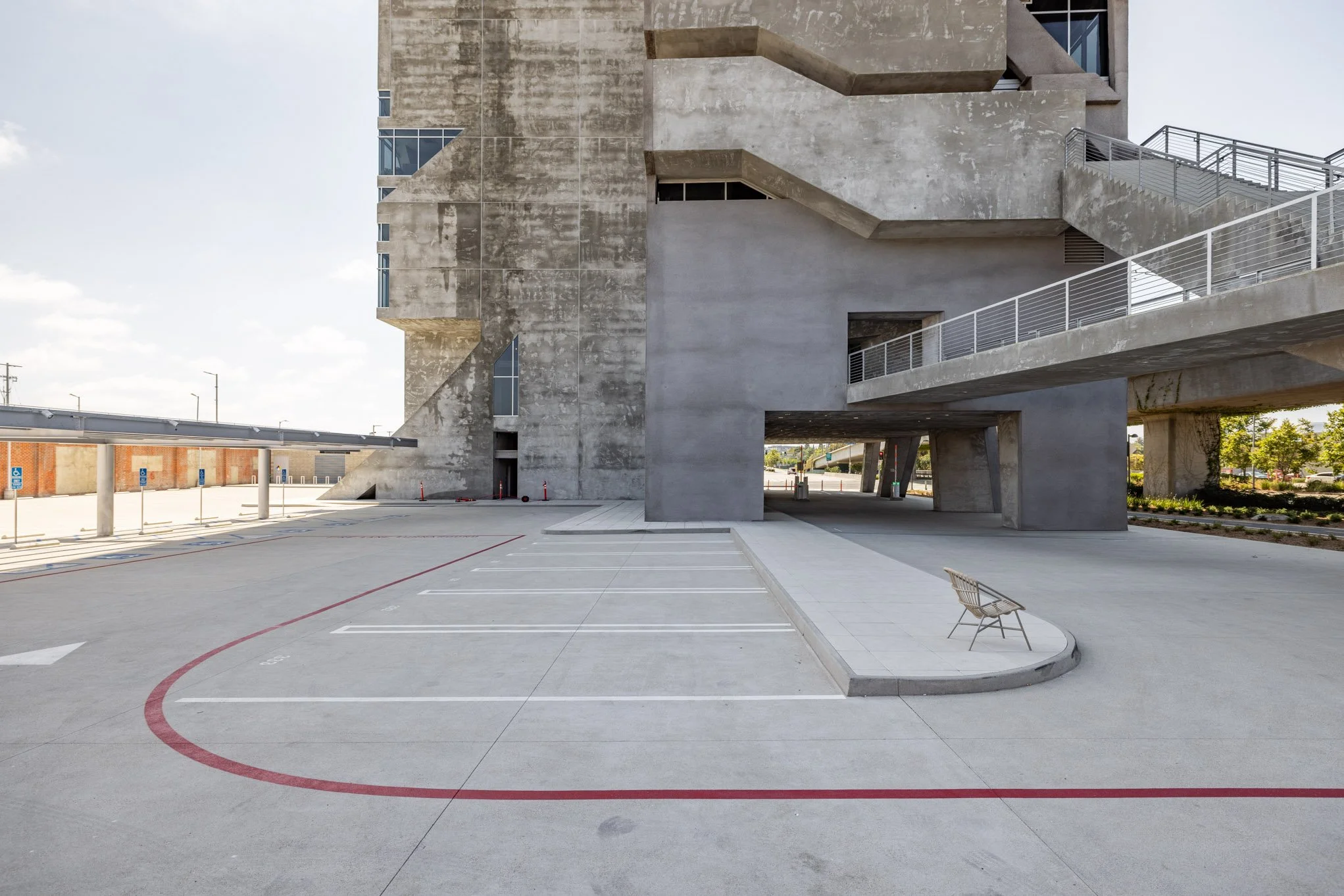 Empty outdoor parking lot with marked parking spaces, a small bench, and a large modern concrete building with stairs and windows.