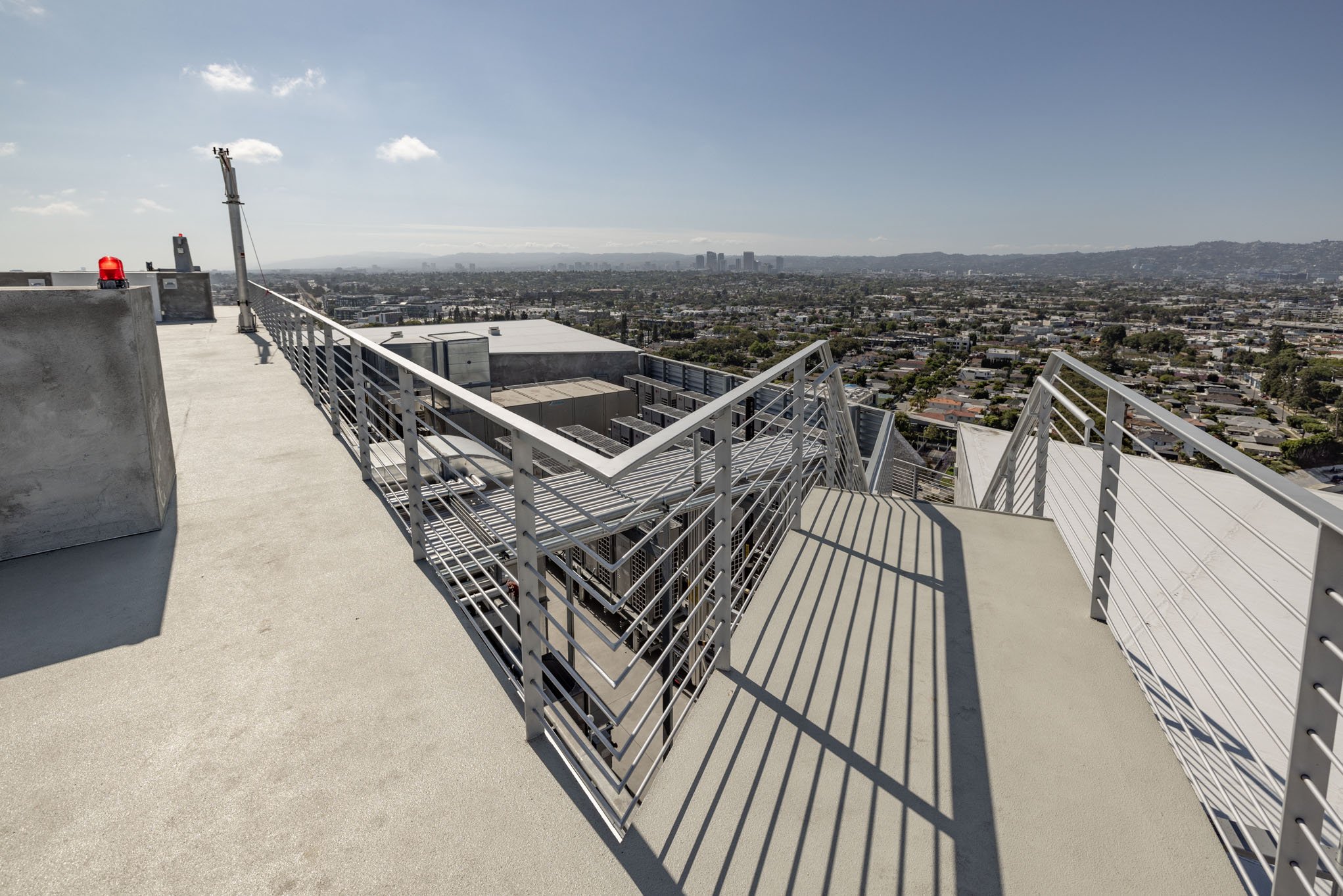 View from the rooftop of a high-rise building showing metal railings, HVAC units below, and a cityscape in the distance under a partly cloudy sky.