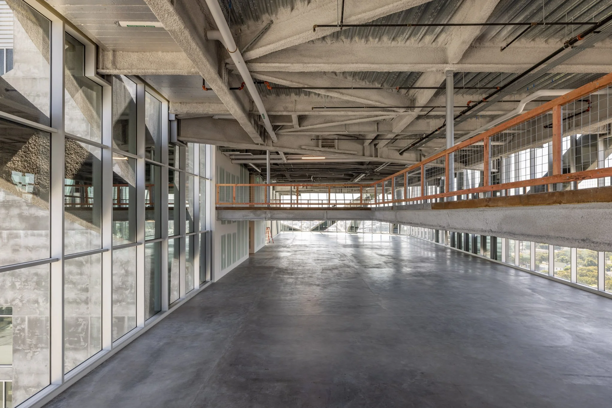 Interior of a modern building under construction with large floor-to-ceiling windows, exposed concrete ceiling and floor, and a suspended walkway with wooden railings.
