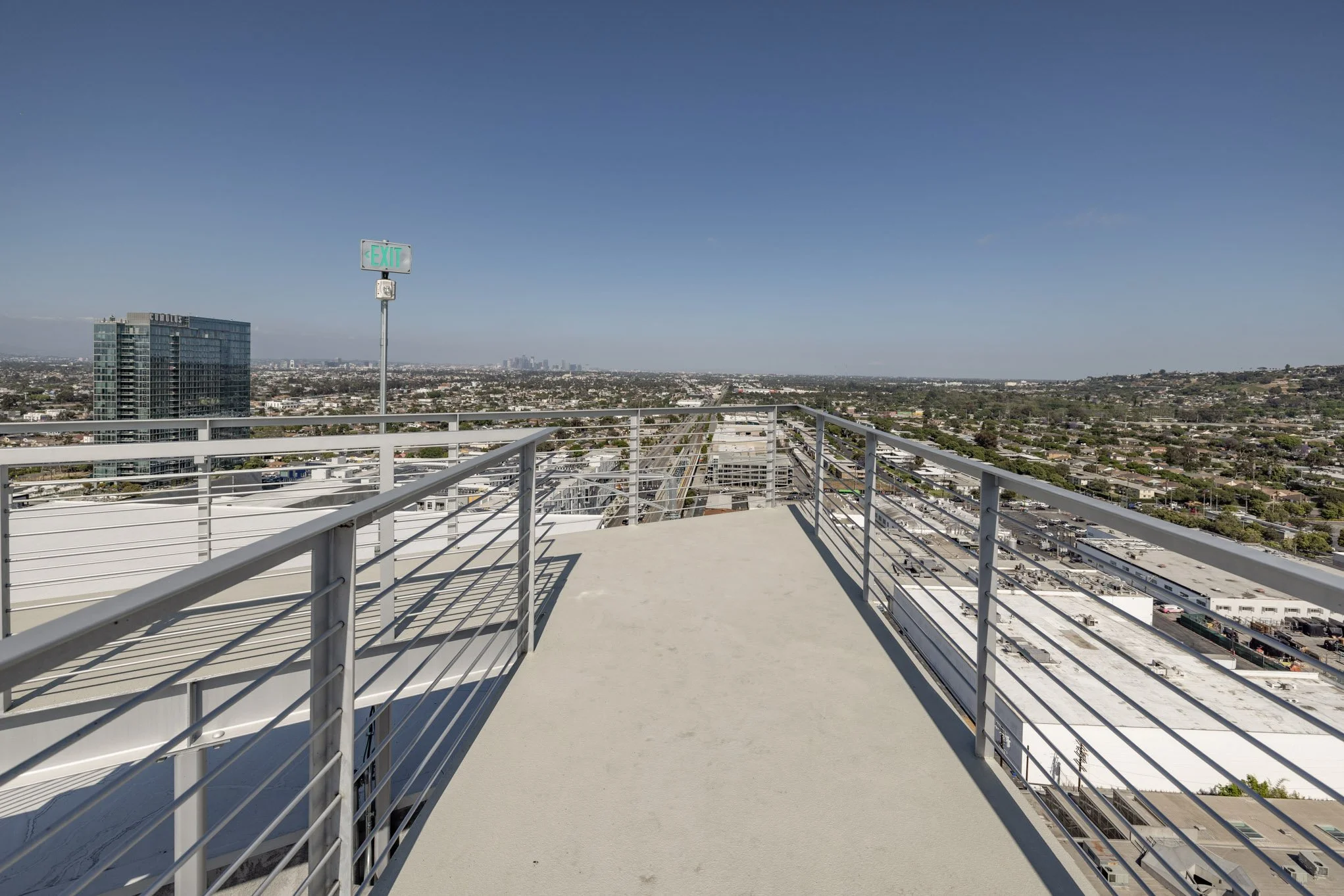 A rooftop terrace with metal railing overlooking a cityscape and distant skyline under a blue sky.