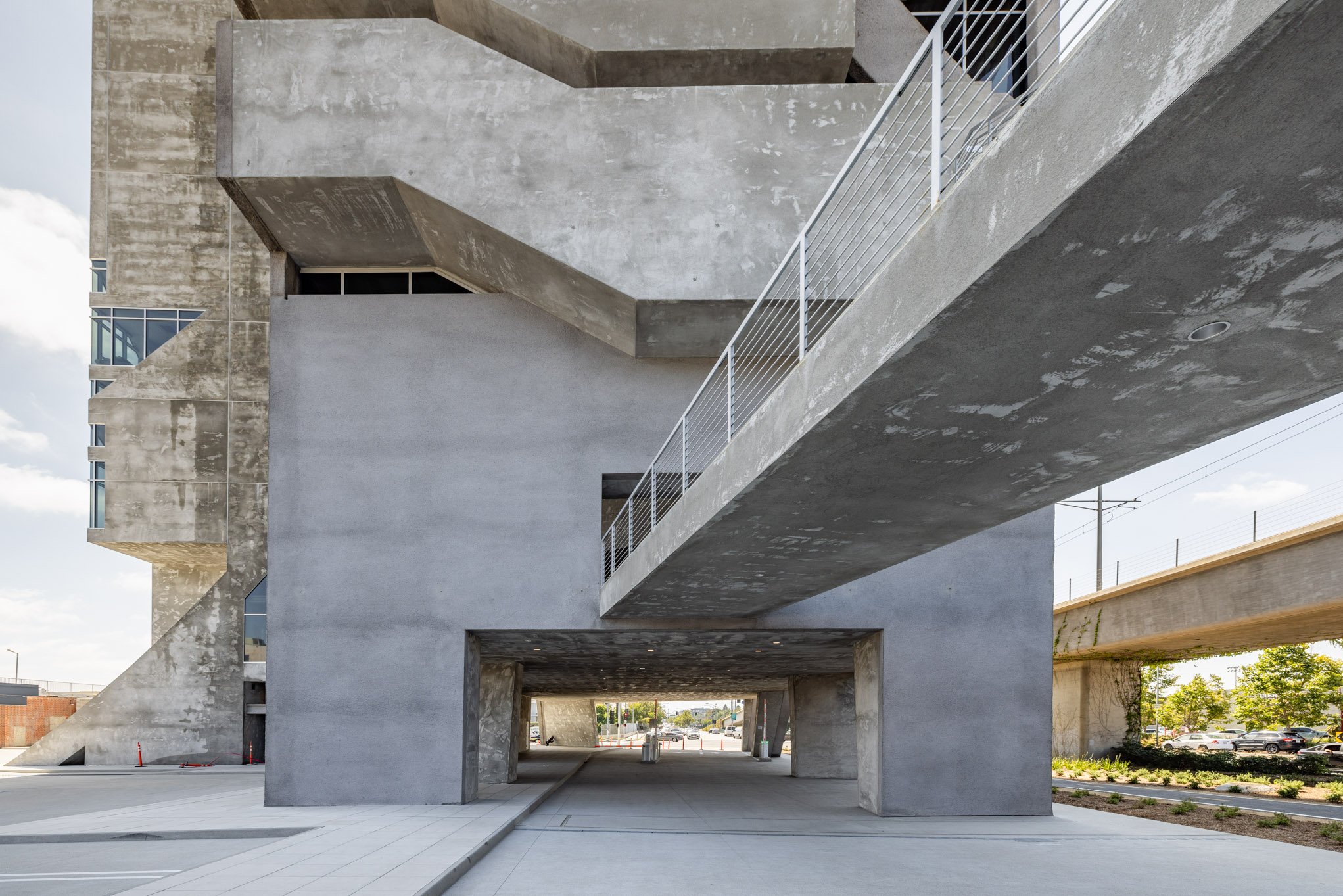 Modern concrete building with geometric design and walkways, underpass visible with a street and trees in the background.