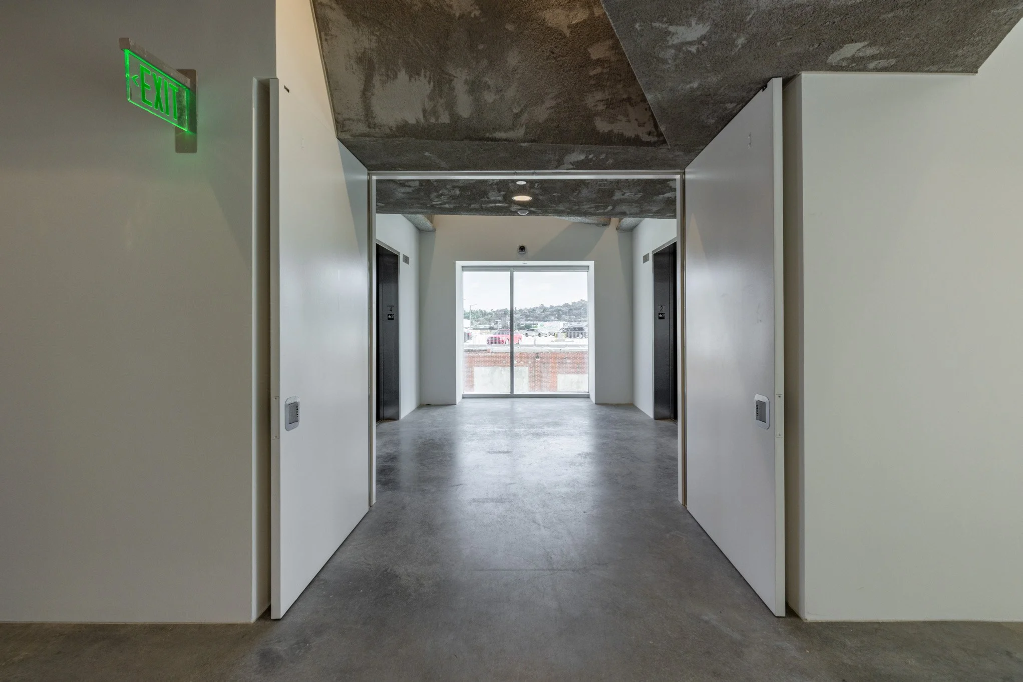 Corridor with two elevator doors on the sides and a large window at the end, showing a parking lot and distant hills outside.