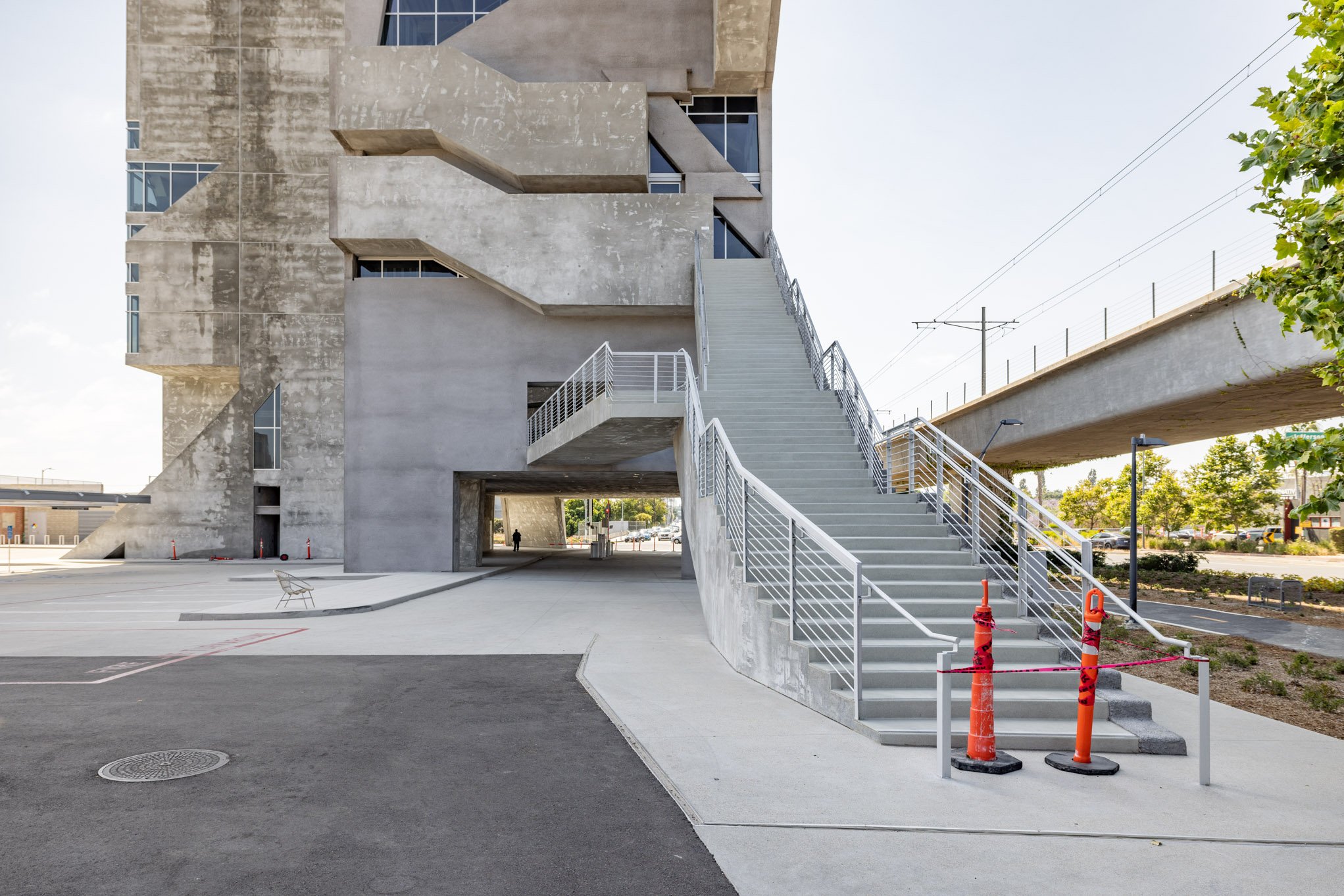 Concrete building with external staircase, elevated train track, and parking area with barriers.