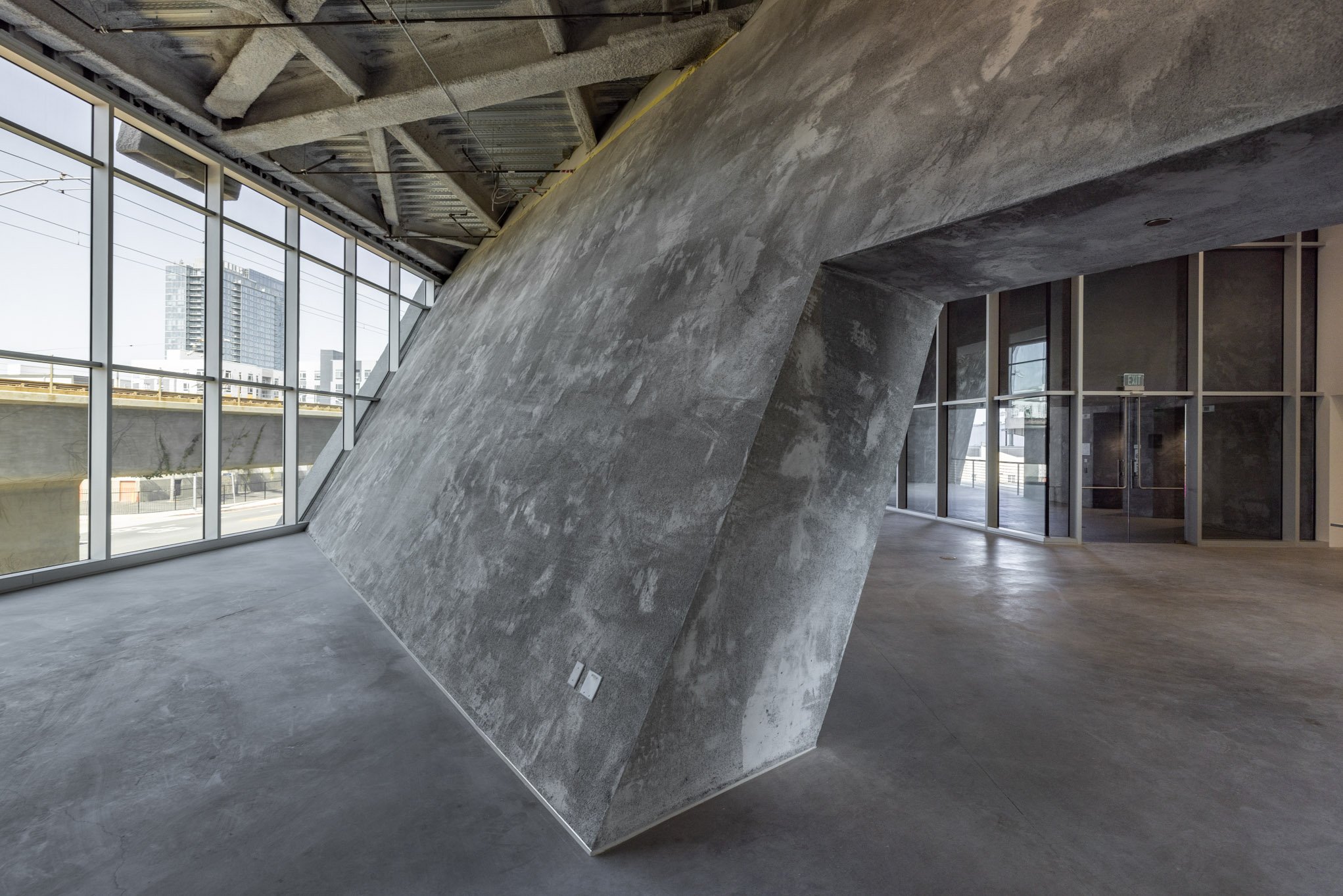 Modern interior space with a large, angular, concrete wall feature, floor-to-ceiling windows, and exposed structural elements.