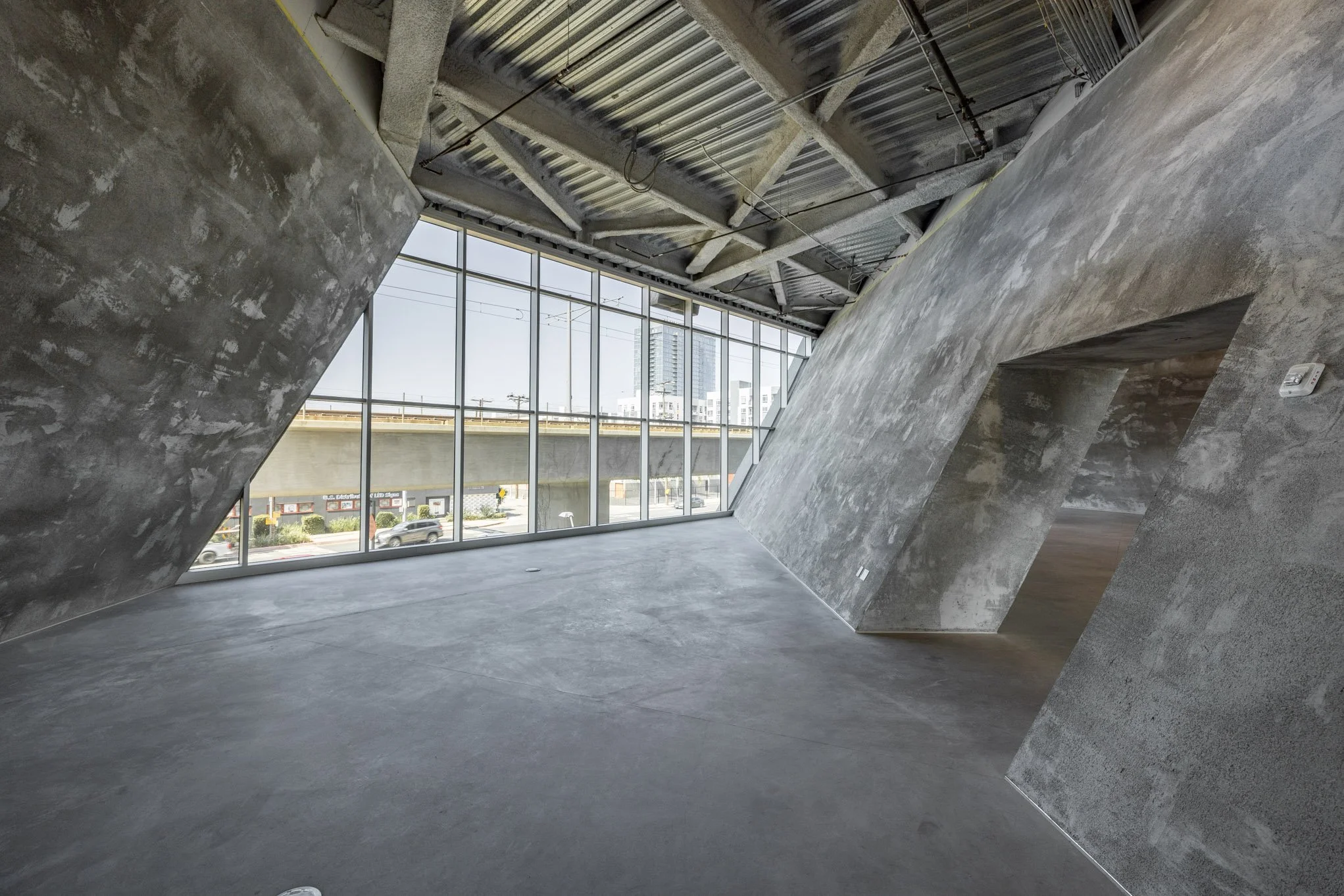 Modern interior space with angular concrete walls and large floor-to-ceiling windows overlooking a street with cars and a highway overpass.