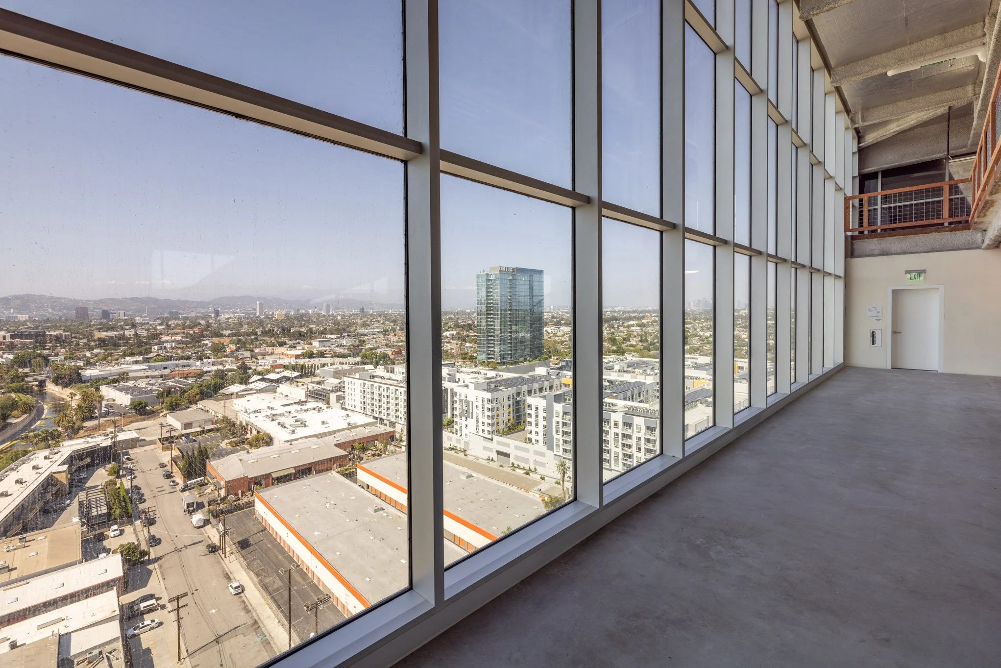 Empty room with large floor-to-ceiling windows overlooking a cityscape with high-rise buildings and streets, under a clear sky.