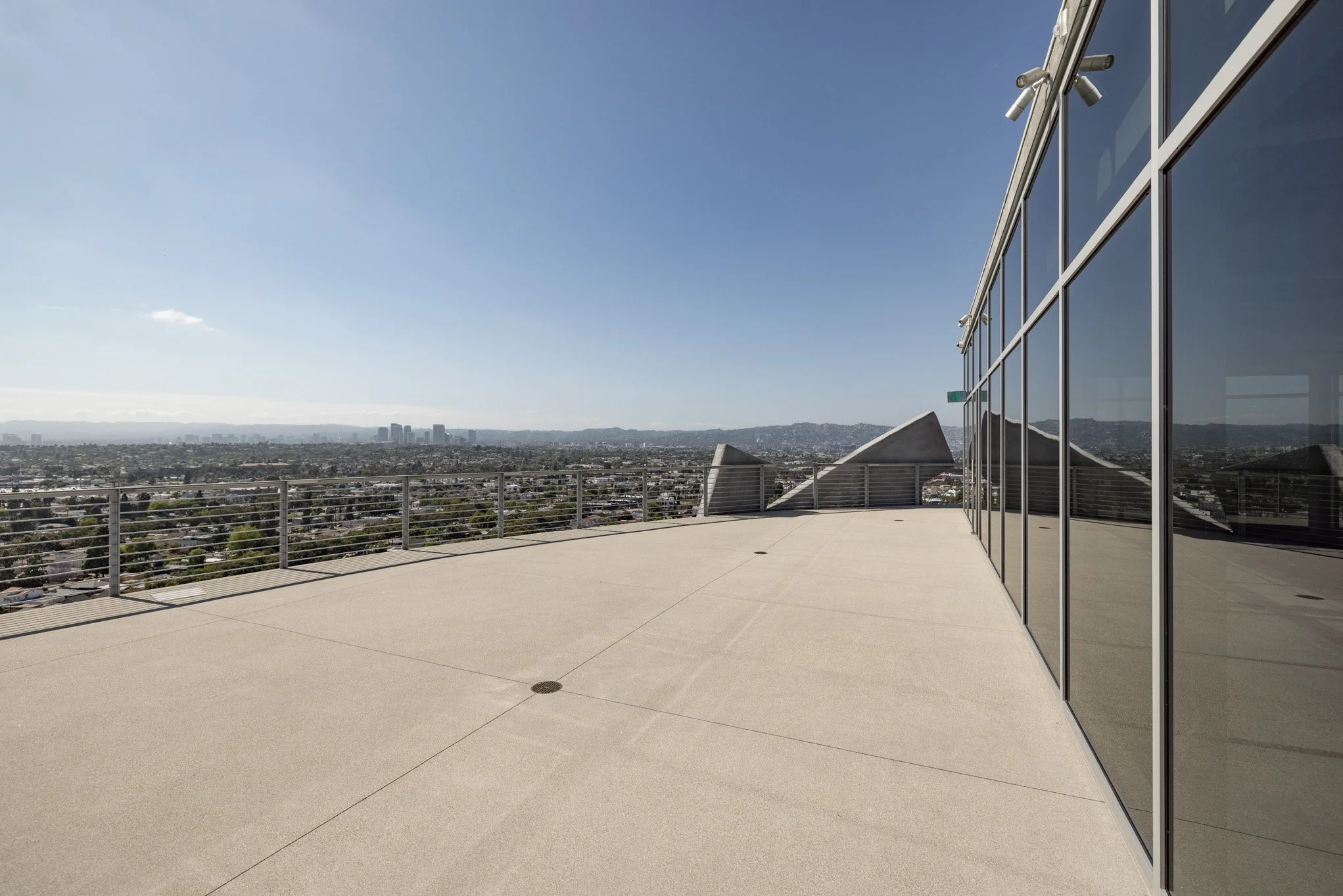 Empty balcony with glass railing and city view in the distance under a clear blue sky.