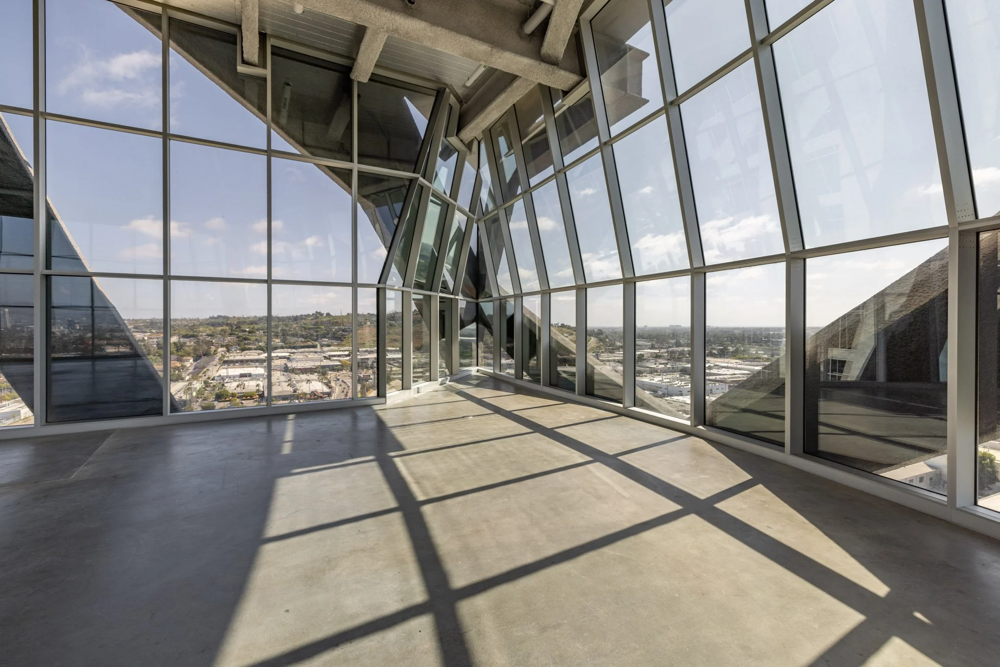 Empty room with large floor-to-ceiling windows showing a cityscape and blue sky outside.