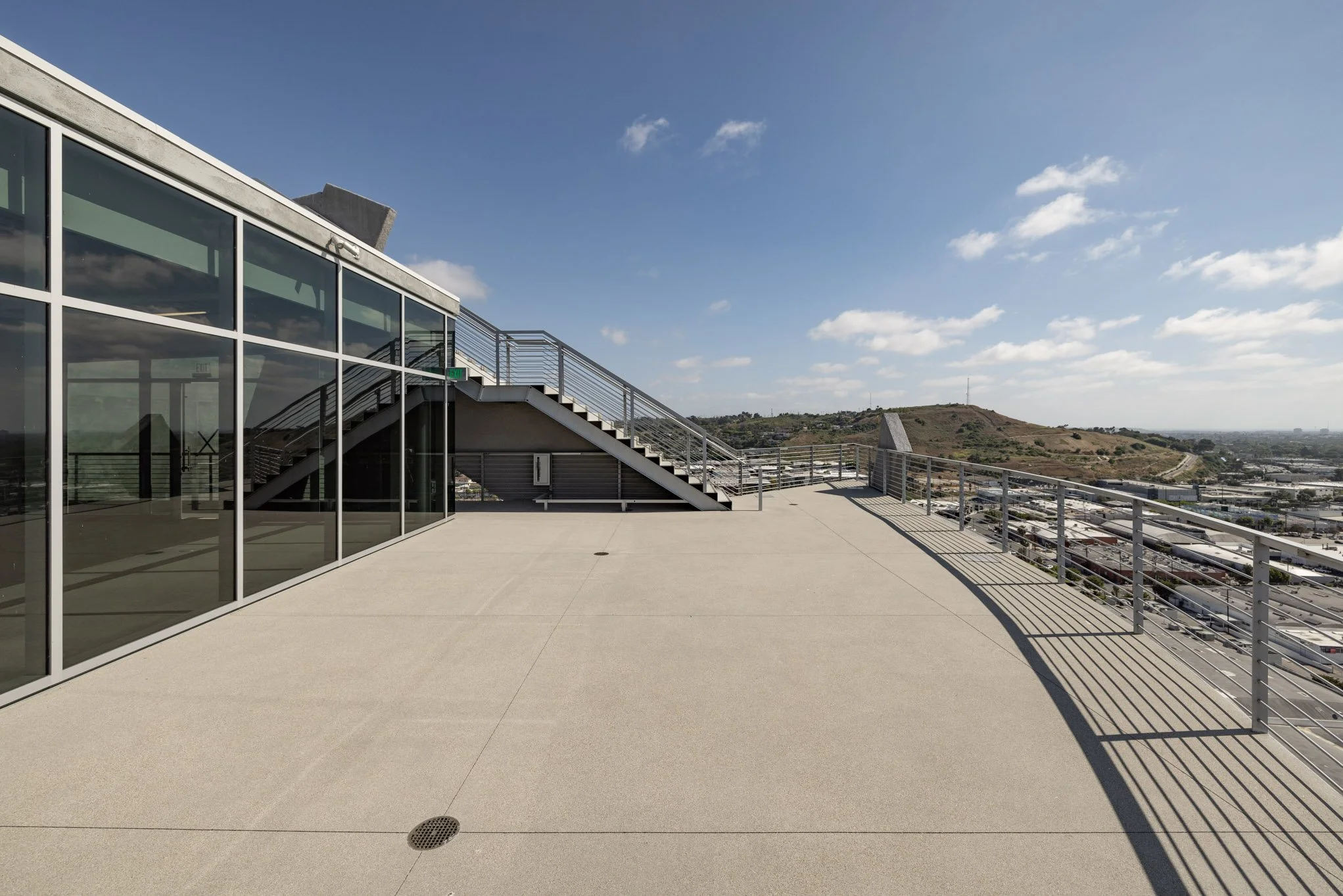 Rooftop terrace with glass walls, metal staircase, and city views under a partly cloudy sky.