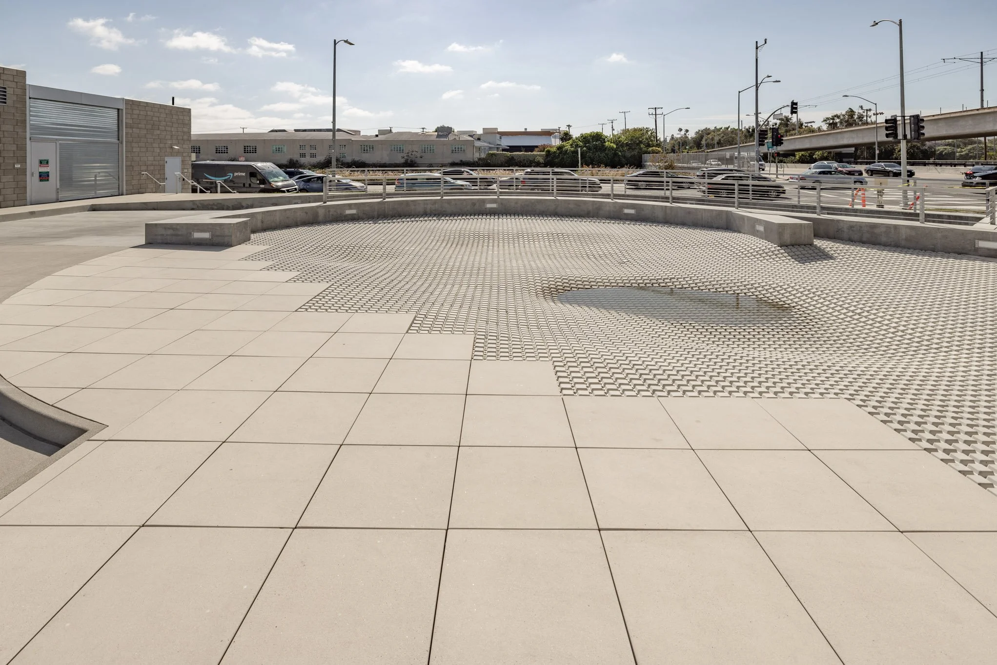 Empty outdoor plaza with a tiled and patterned concrete surface, concrete benches, surrounding street, parked cars, streetlights, traffic lights, and buildings in the background.