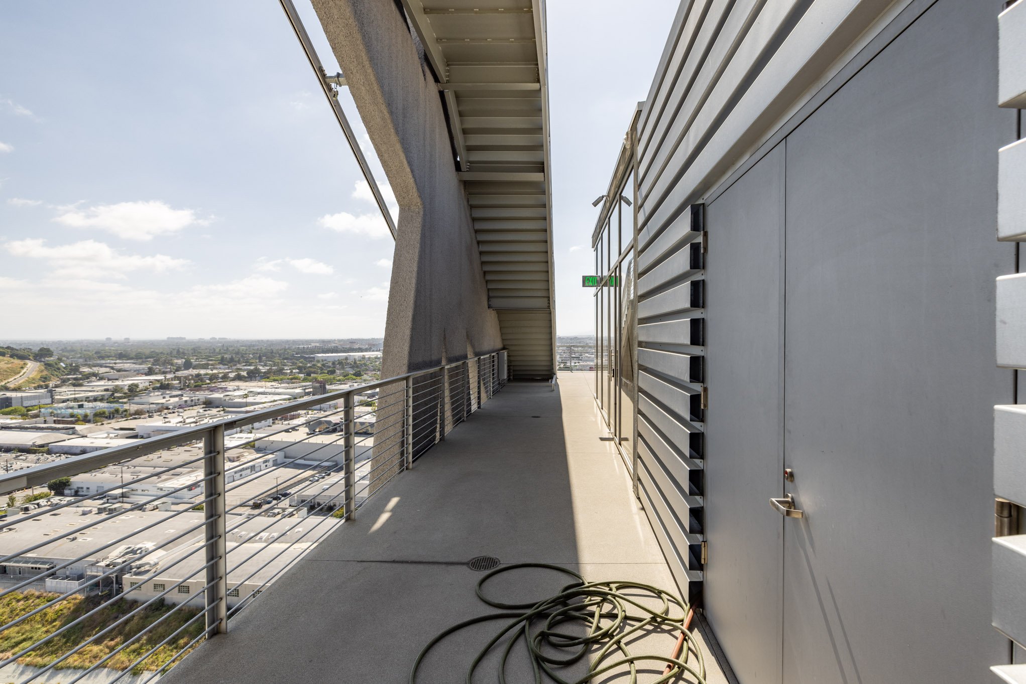 An empty rooftop balcony with a metal railing, outdoor electrical cable, and view of a cityscape and sky in the background.