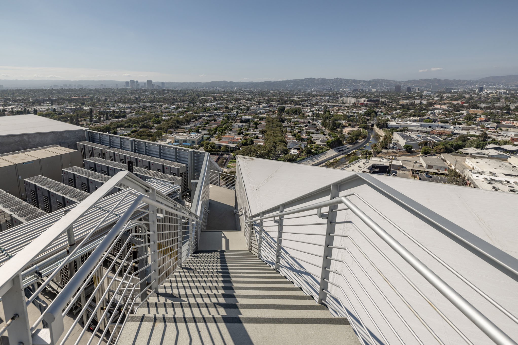 A high rooftop stairway with metal railings overlooks an urban cityscape with houses, roads, and distant high-rise buildings under a clear blue sky.