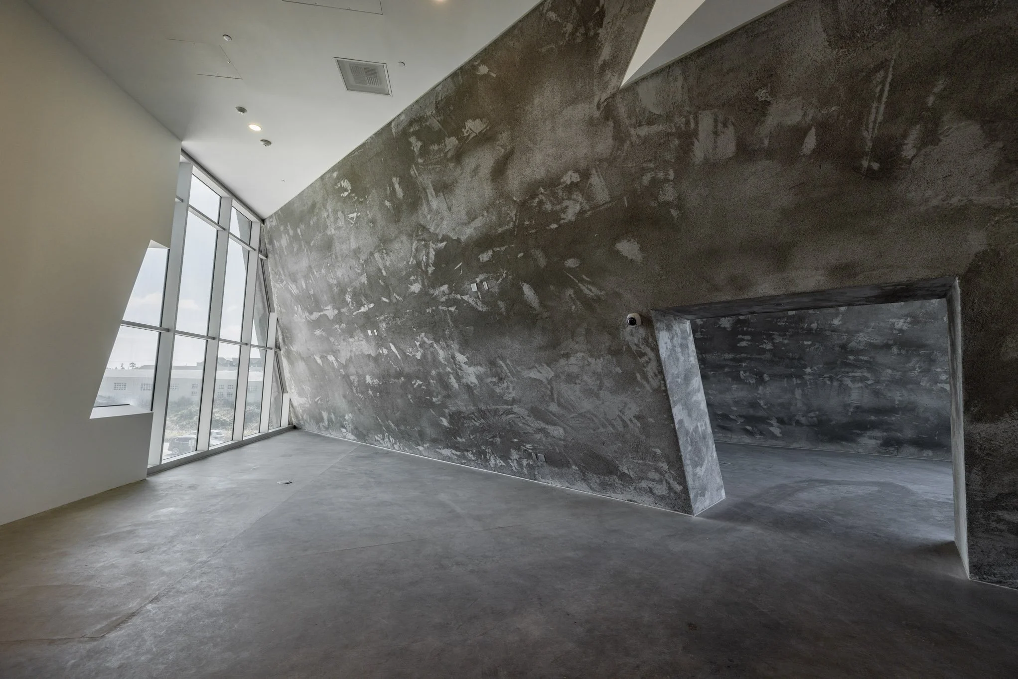 Modern interior space with large windows, unfinished concrete walls, and a concrete floor. The wall on the right side features a large rectangular opening, and the ceiling has recessed lighting.