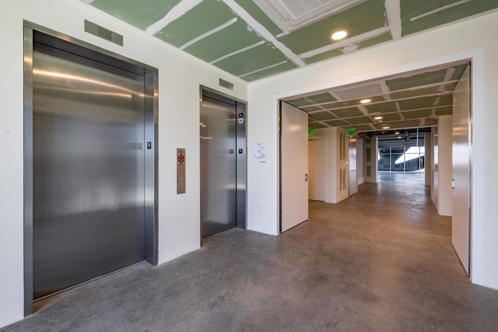 An interior scene showing two closed elevator doors made of stainless steel, with a hallway extending beyond, in a building under construction or renovation.