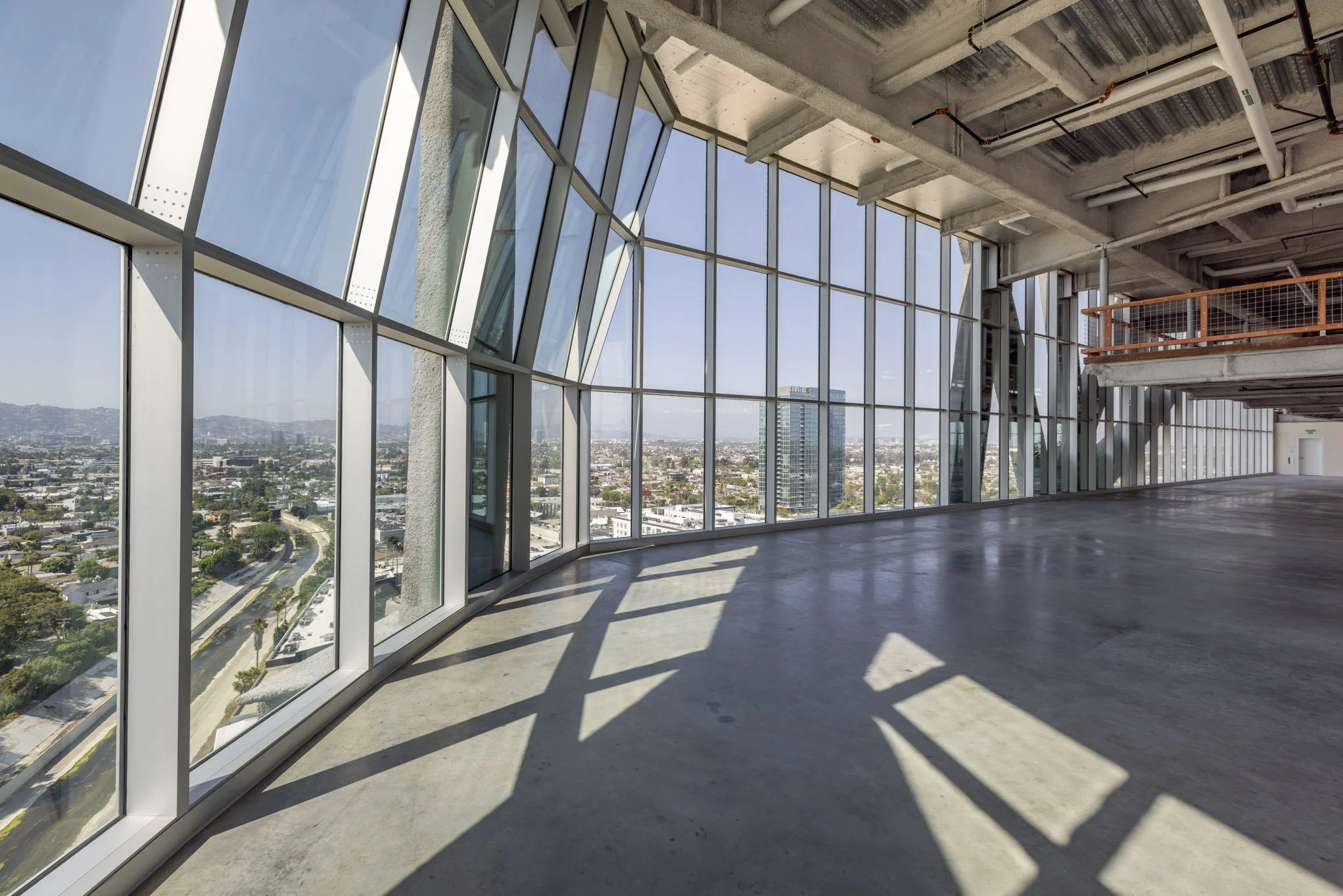 Interior of an unfinished building with large glass windows, concrete floors, and exposed ceiling beams, with cityscape view outside.