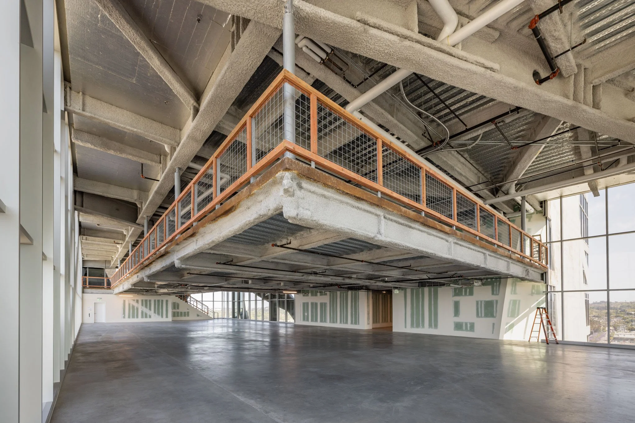 Empty indoors construction site with an unfinished second floor, concrete floors, visible piping and ductwork, large windows, and a ladder.