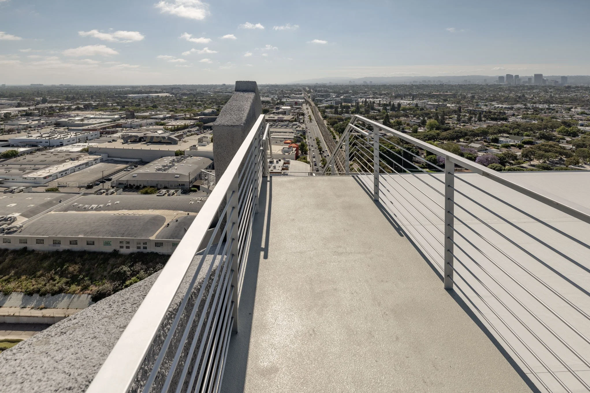 View from a rooftop or high balcony showing cityscape and clear sky with some clouds, metal railings on the rooftop edge.