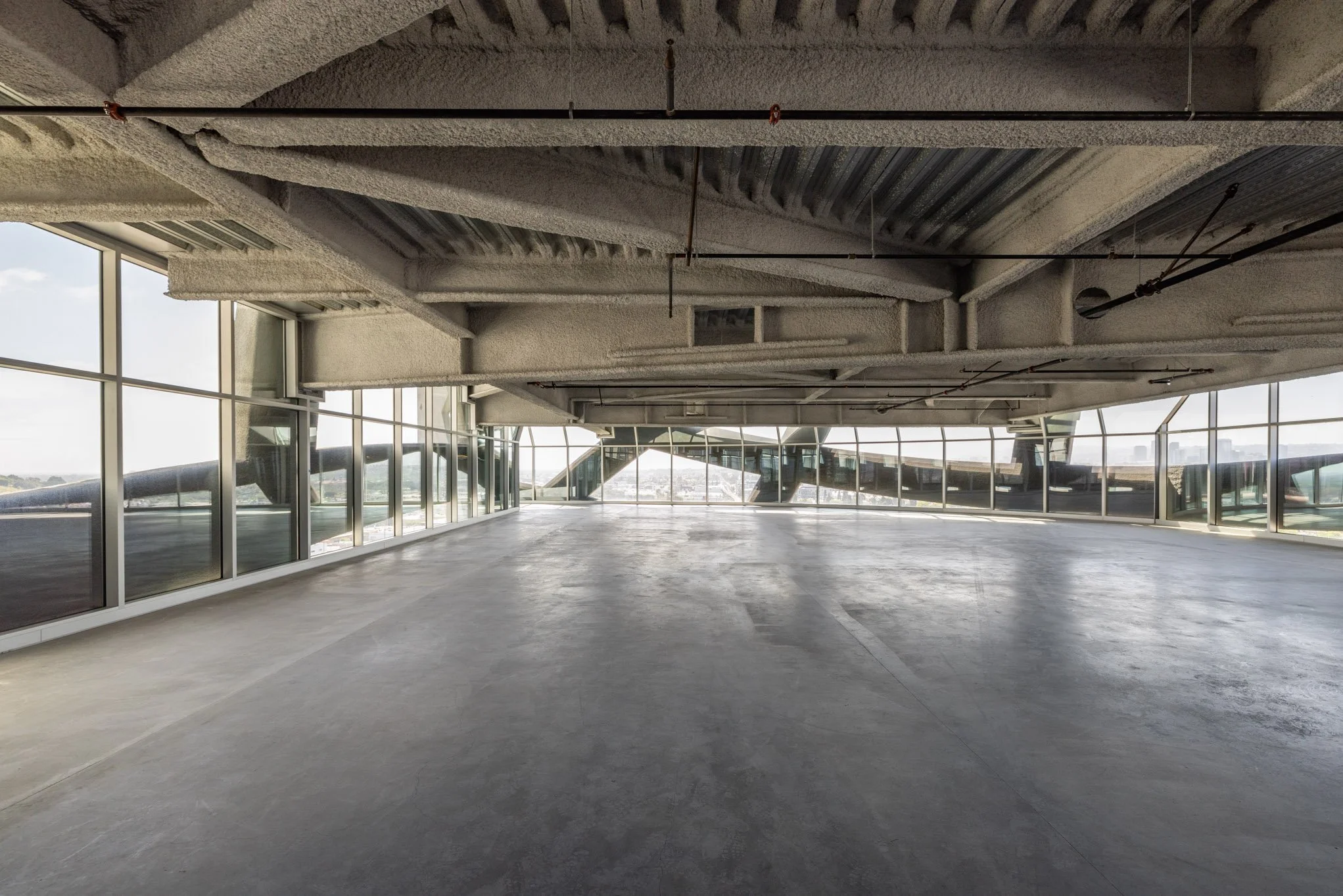 Empty, open indoor space with large glass windows, concrete floor, and exposed ceiling beams.