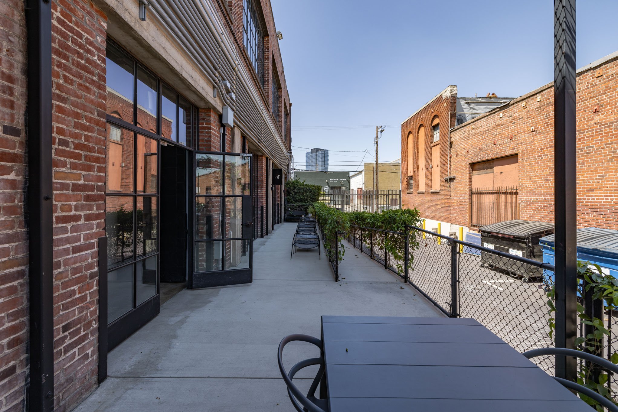 Urban outdoor patio with black metal table and chairs, brick building on the left and brick wall on the right, sidewalk with trash bins and trash can, city skyscraper visible in the distance under a clear blue sky.